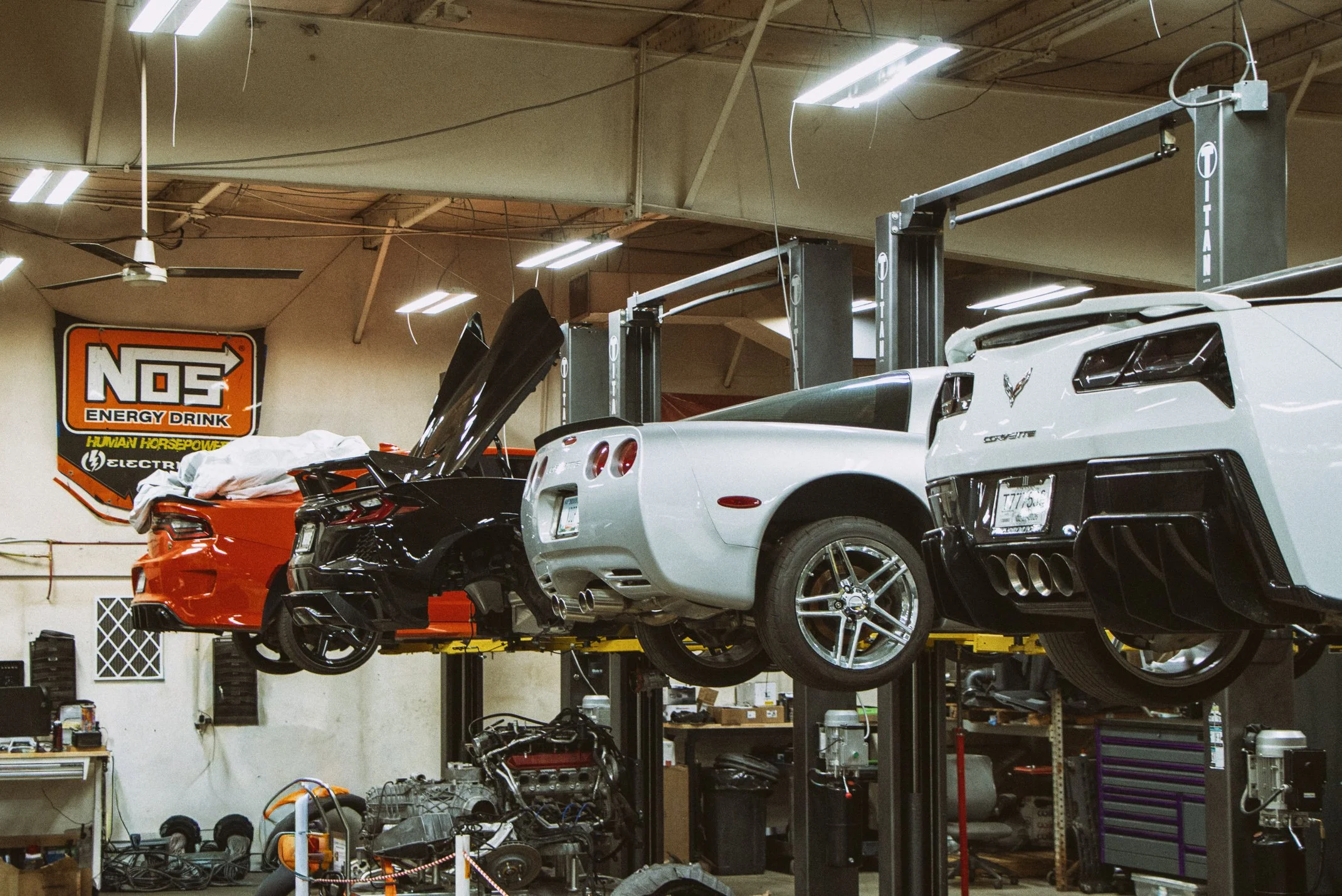 A garage or workshop with two sports cars parked on hydraulic lifts, one red and one white, with various tools and equipment visible in the background.
