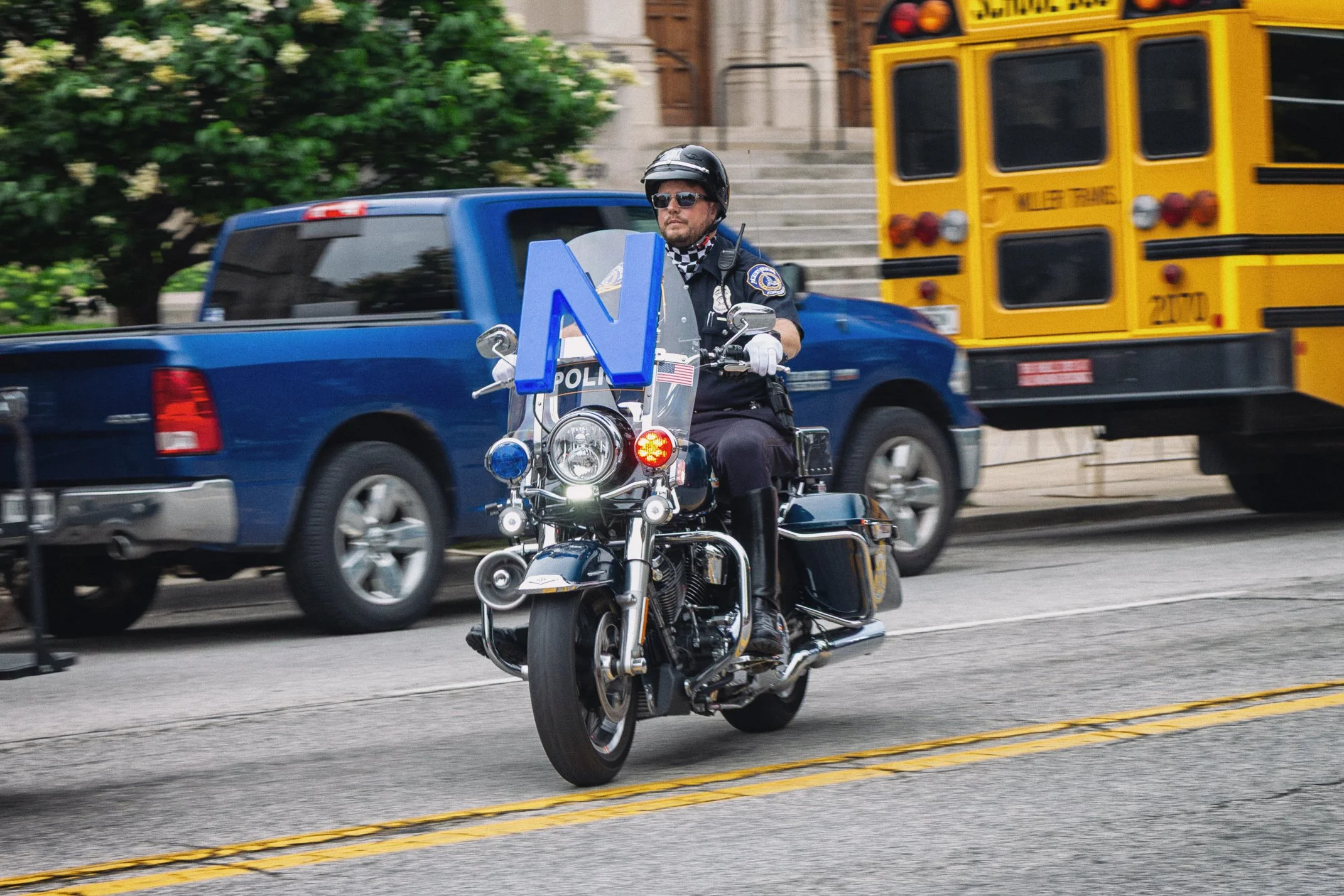 A police officer riding a motorcycle on the street with a yellow school bus and blue truck in the background.