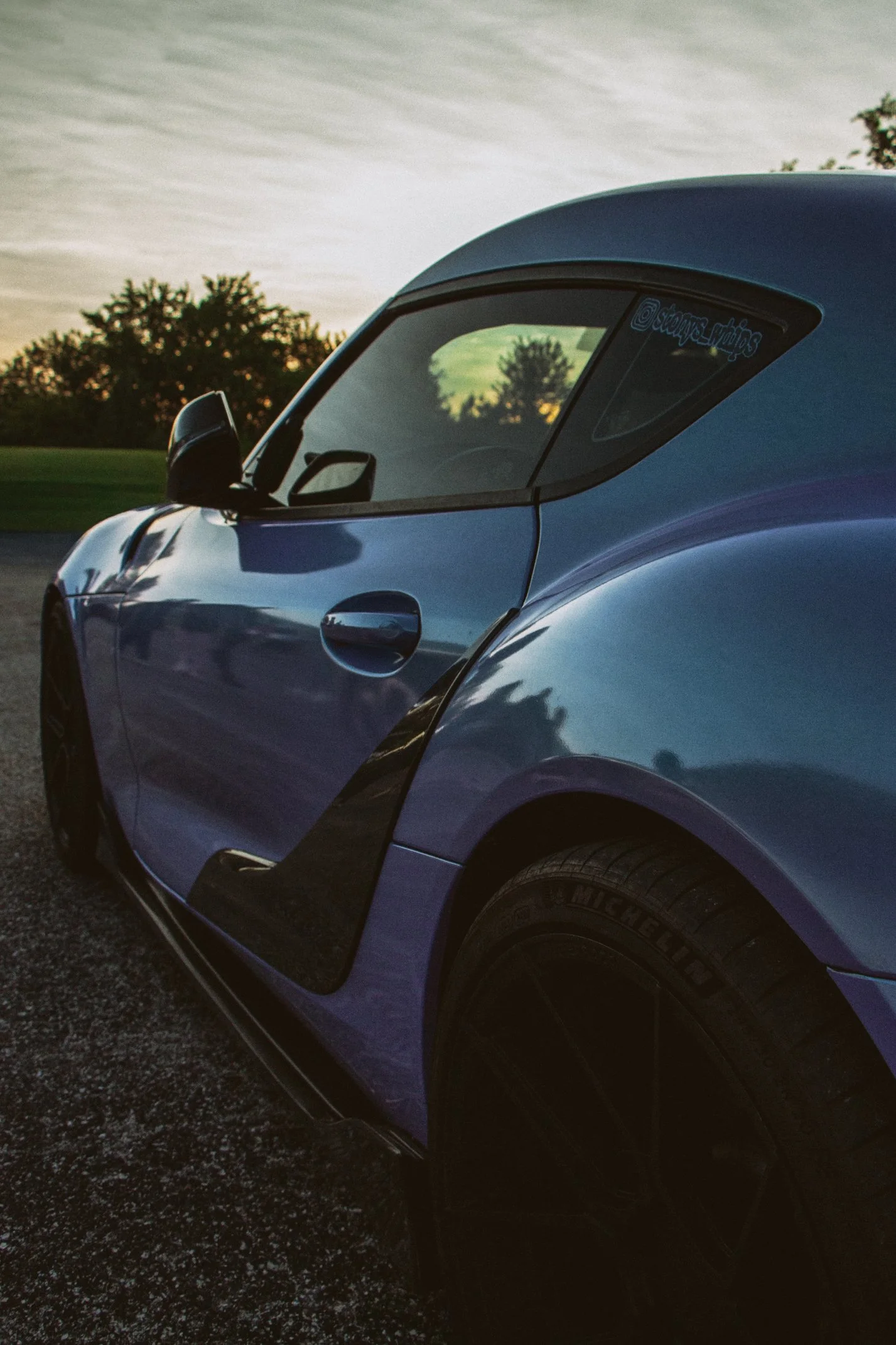 Close-up of a blue sports car parked on a gravel surface during sunset, with trees and a cloudy sky in the background.