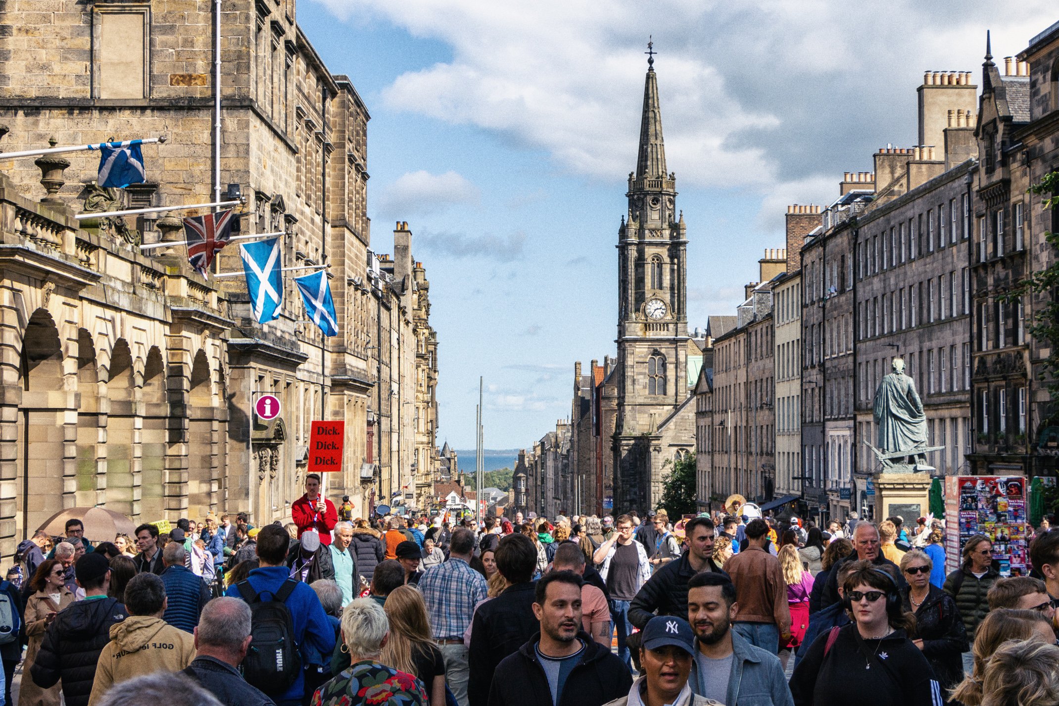 Crowded street in a historic city with churches, flags, and a statue, under a partly cloudy sky.