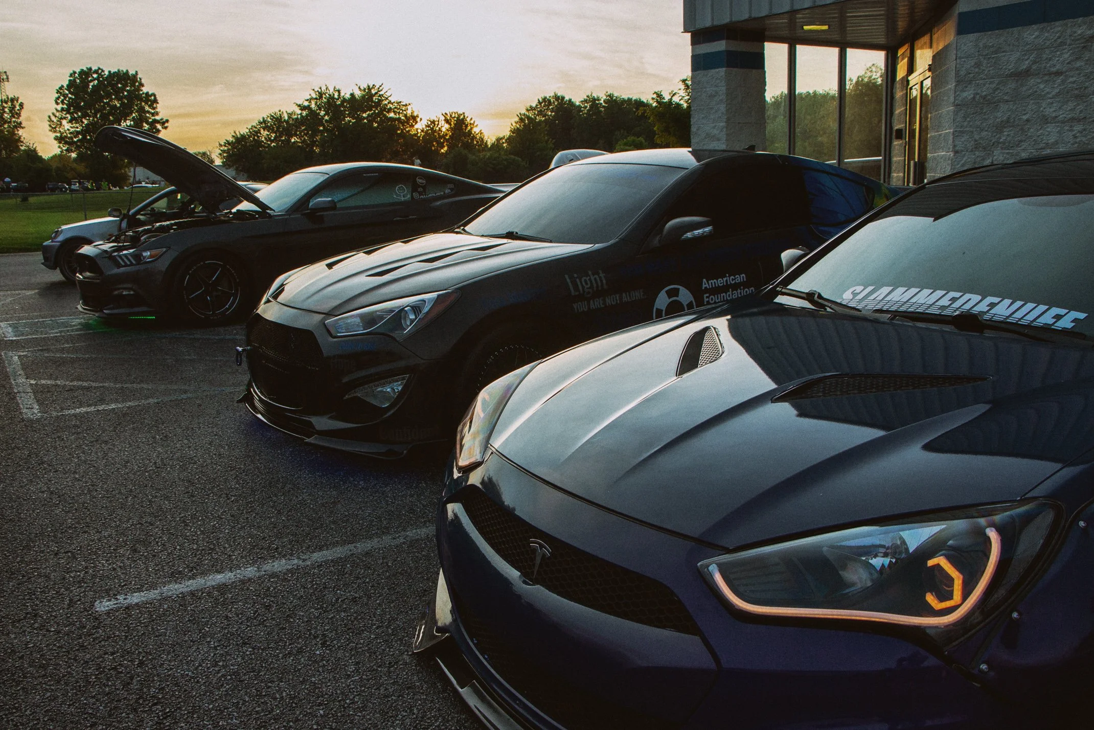 A row of black and gray Tesla cars parked outside a building during sunset, with trees and a cloudy sky in the background.