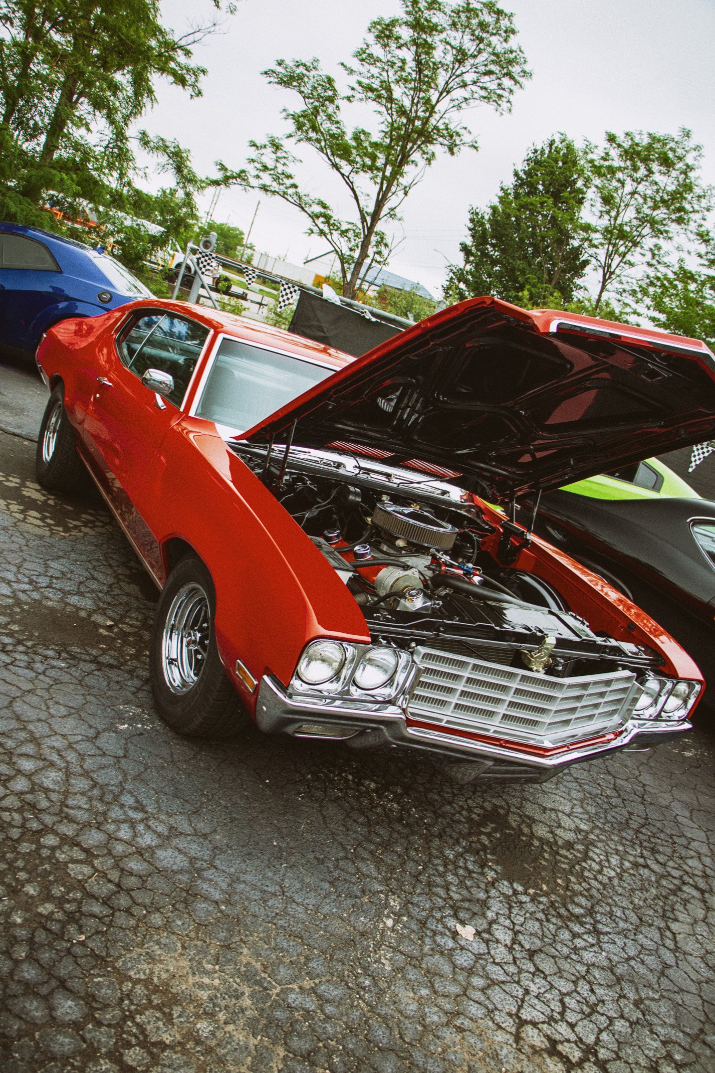 A vintage red car with its hood open, displaying the engine, parked on a cracked asphalt surface alongside other cars at a car show. Trees and flags are visible in the background.
