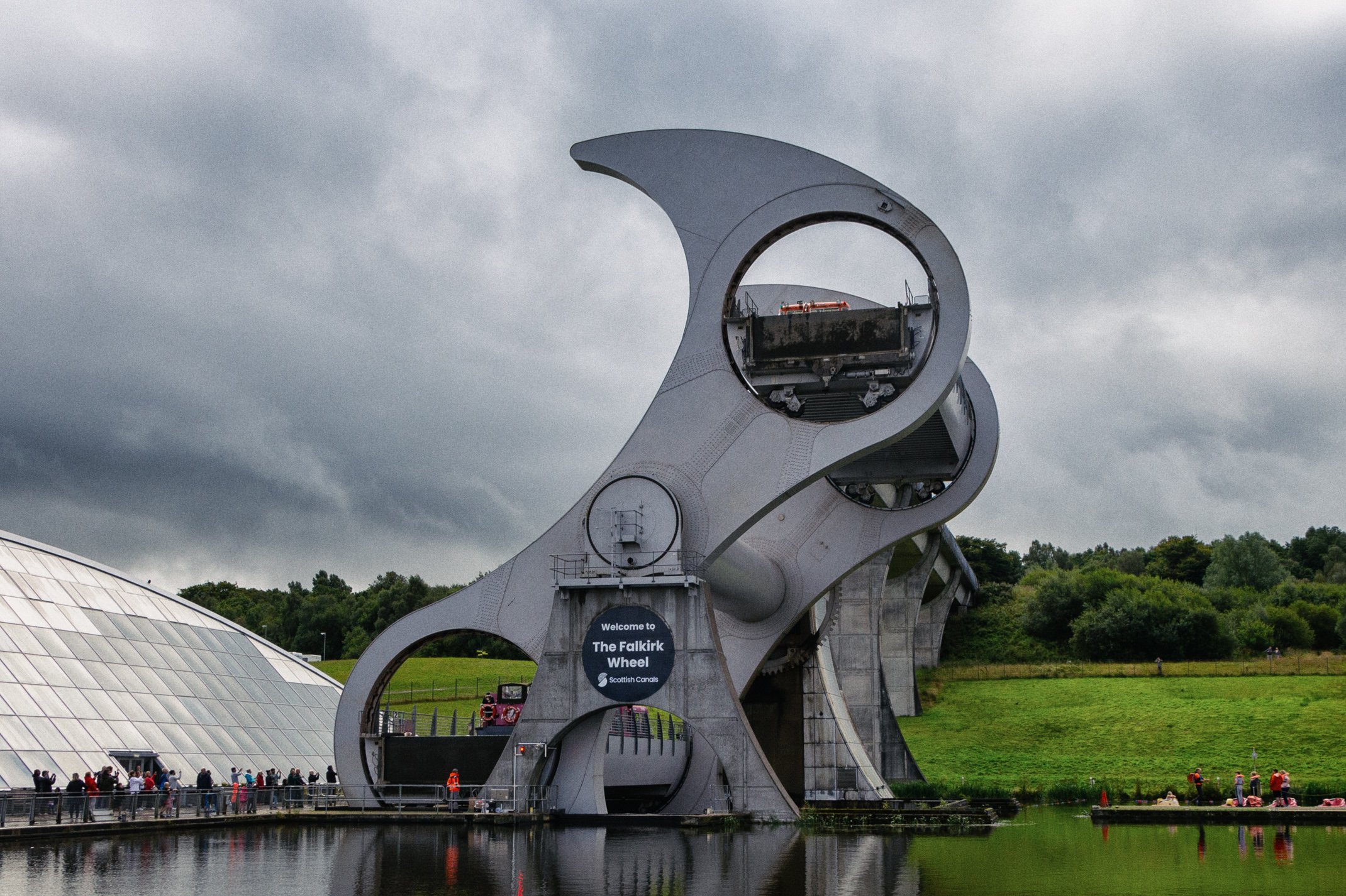 The Falkirk Wheel, a large rotating boat lift in Scotland, with a cloudy sky and visitors standing on the viewing platform.