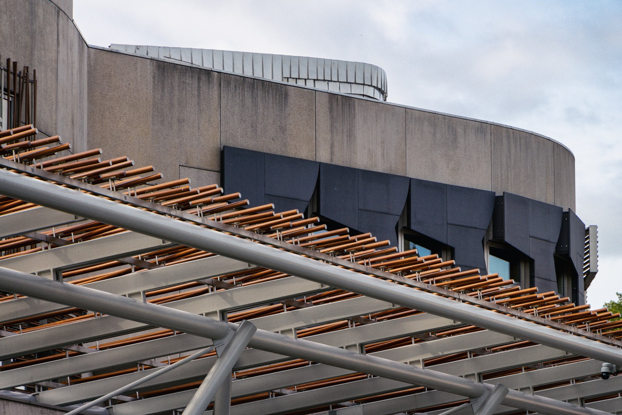 Modern building with geometric black exterior panels and orange sunshade lattice above architectural beams, under cloudy sky.