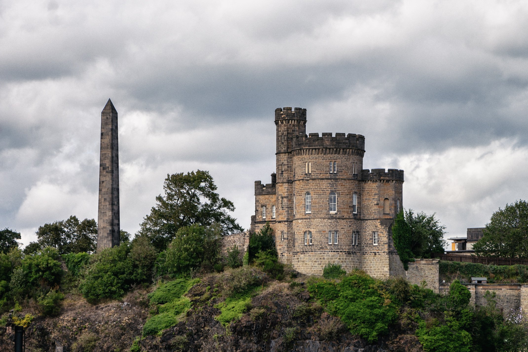 A historic castle with rounded towers and battlements, situated on a hill covered in greenery, with a tall, pointed stone obelisk monument nearby, and a background of cloudy sky.