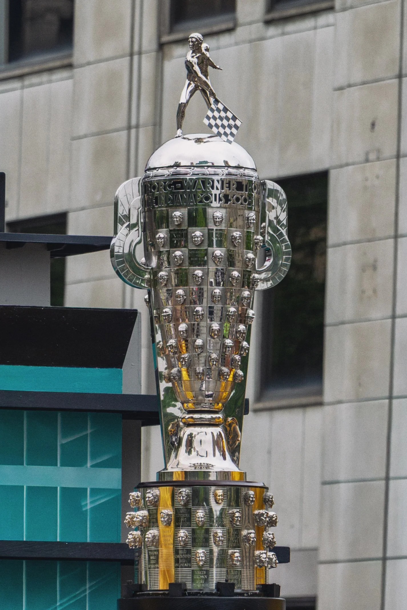 The image shows a large, shiny trophy with a figure of a person on top holding a checkered flag. The trophy has multiple heads of a lion's face around its body and the background features a modern building with windows.