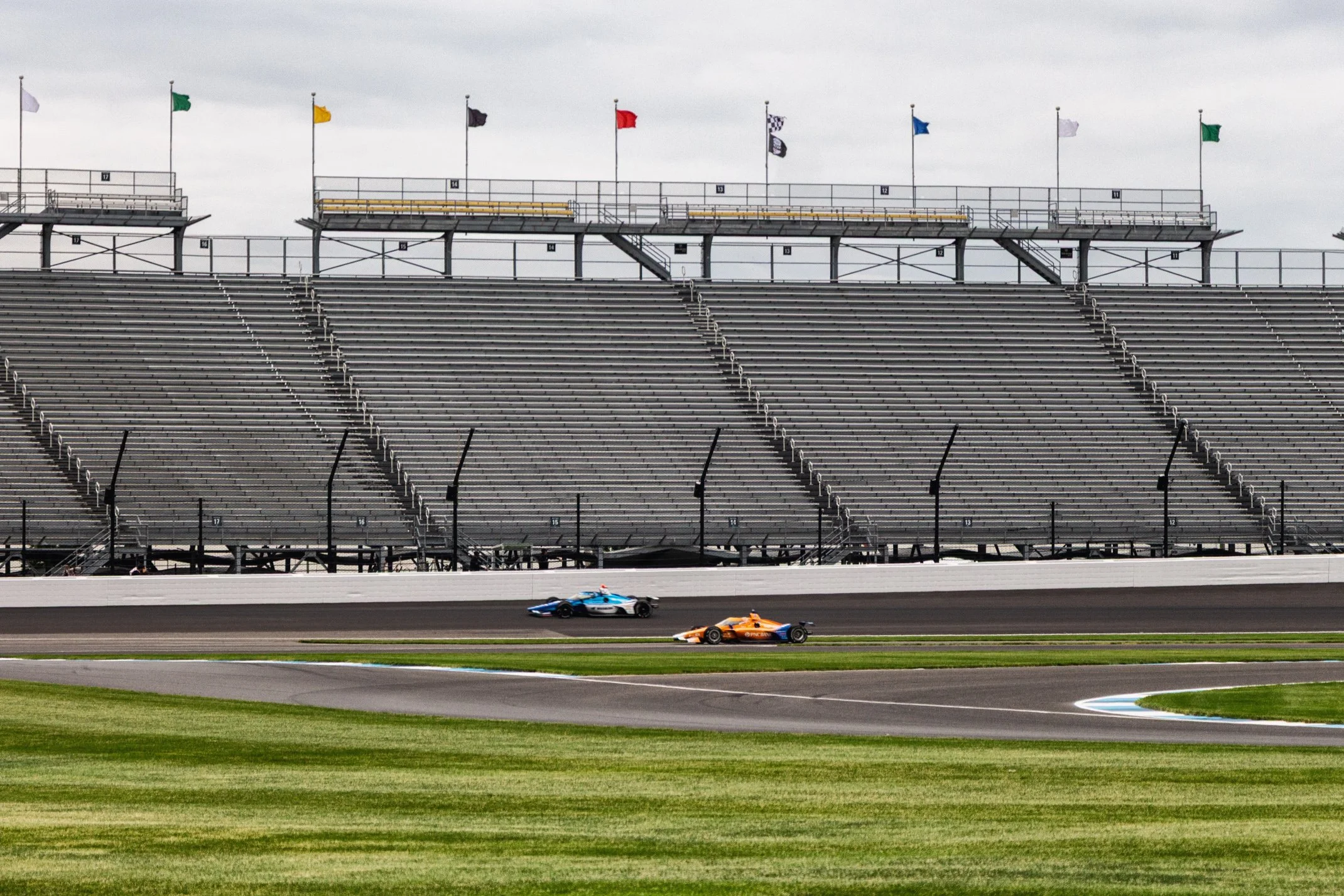 Two race cars speeding on a racing track in front of empty stadium bleachers with multiple flag banners on poles in the background.