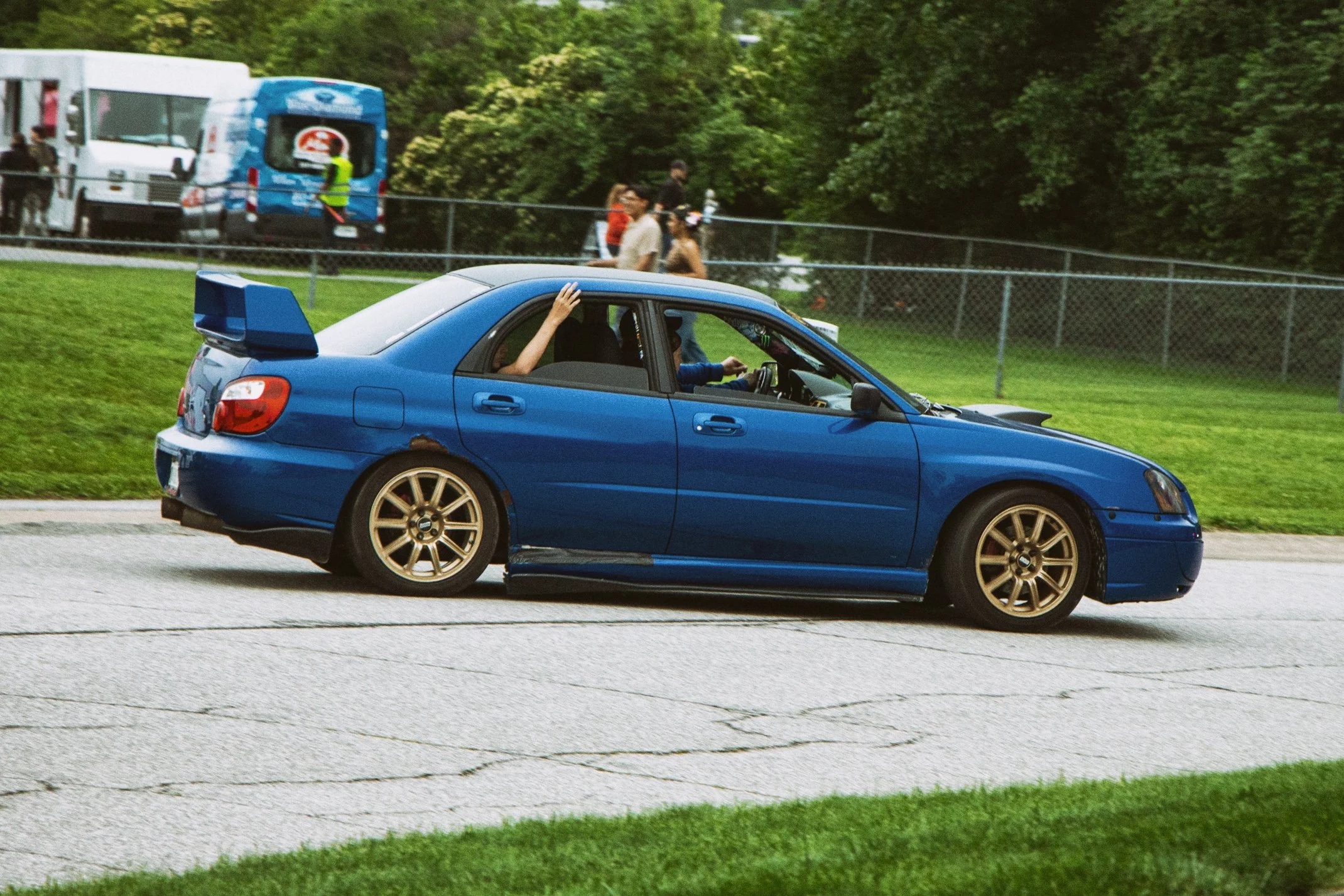 A blue car driving on a road, with a person inside waving their hand from the window. In the background, there are people walking near a chain-link fence and some vehicles parked, surrounded by green trees.
