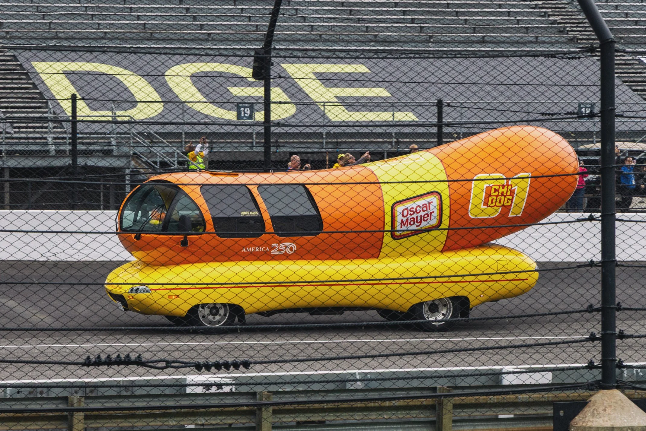 A hot dog shaped vehicle painted in bright yellow with orange and yellow accents, featuring advertisers for Oscar Mayer and Chidog, parked on a race track with spectators and photographers behind a fence in the background.