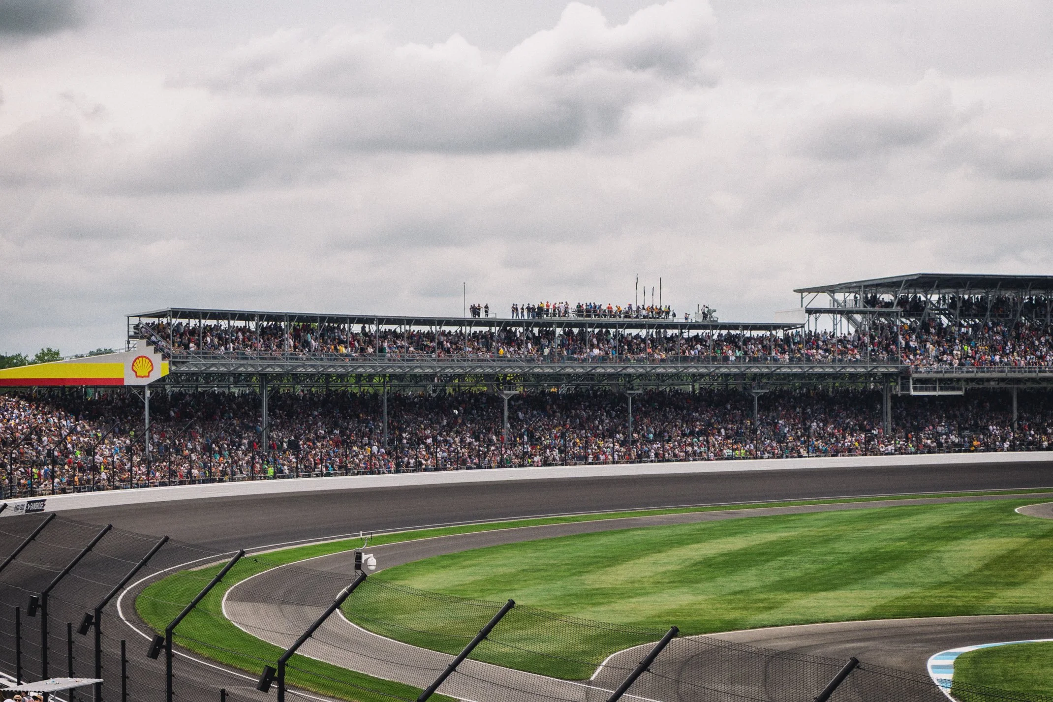 A racing stadium filled with spectators, featuring multiple tiers of seating under cloudy skies, and a section with a Shell logo on a building to the left.