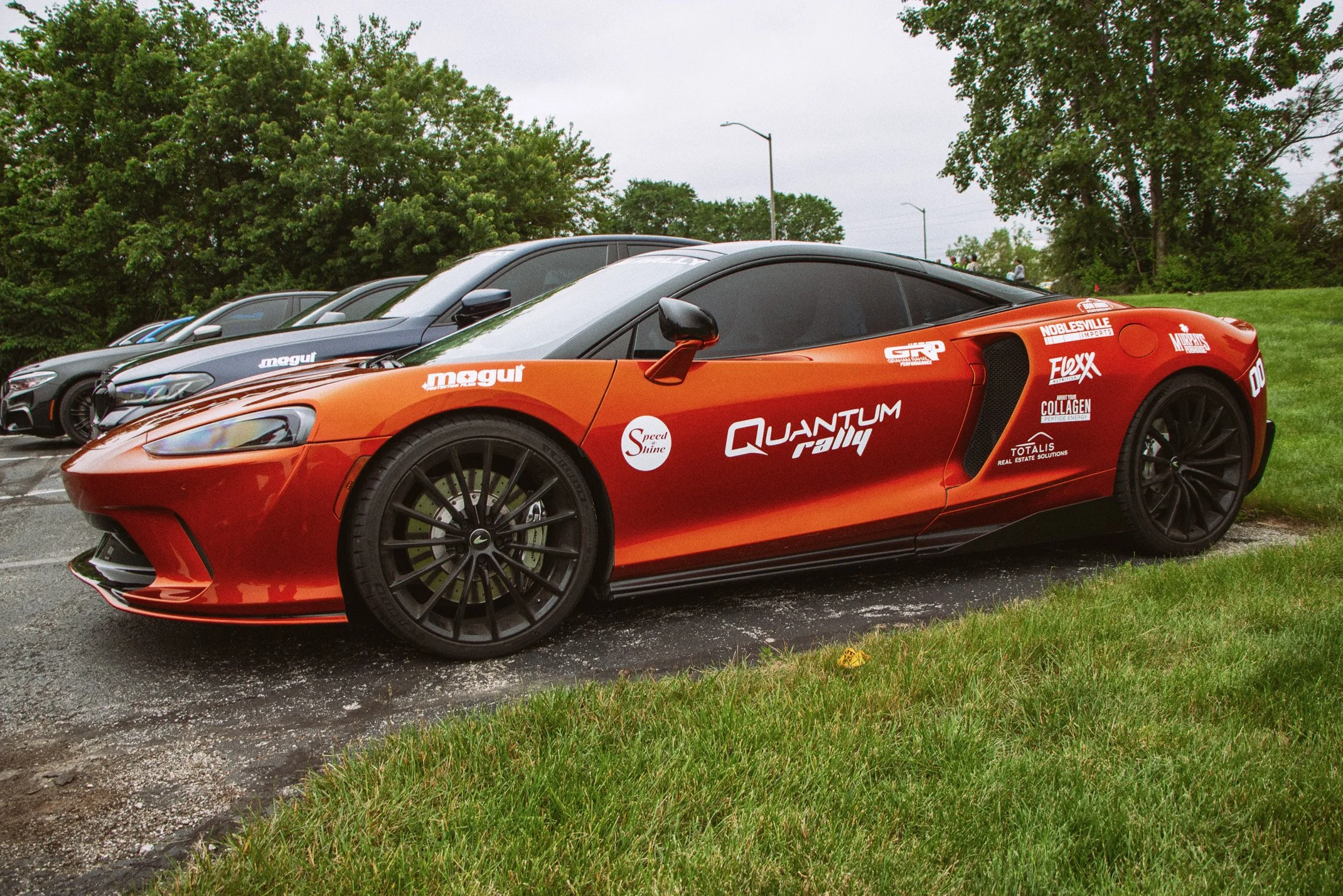 A row of sports cars parked on a grassy area with trees and streetlights in the background, featuring a bright orange car with branding and sponsorship decals in the foreground.