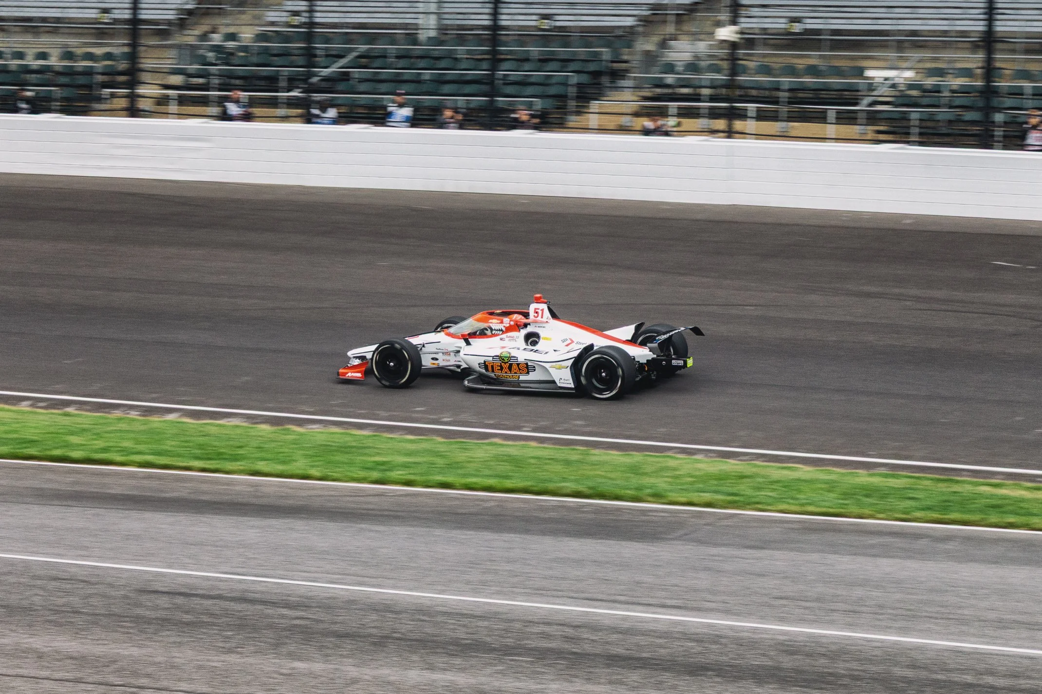 A race car with a red, white, and black design speeding on a racetrack, with blurred spectators and stands in the background.