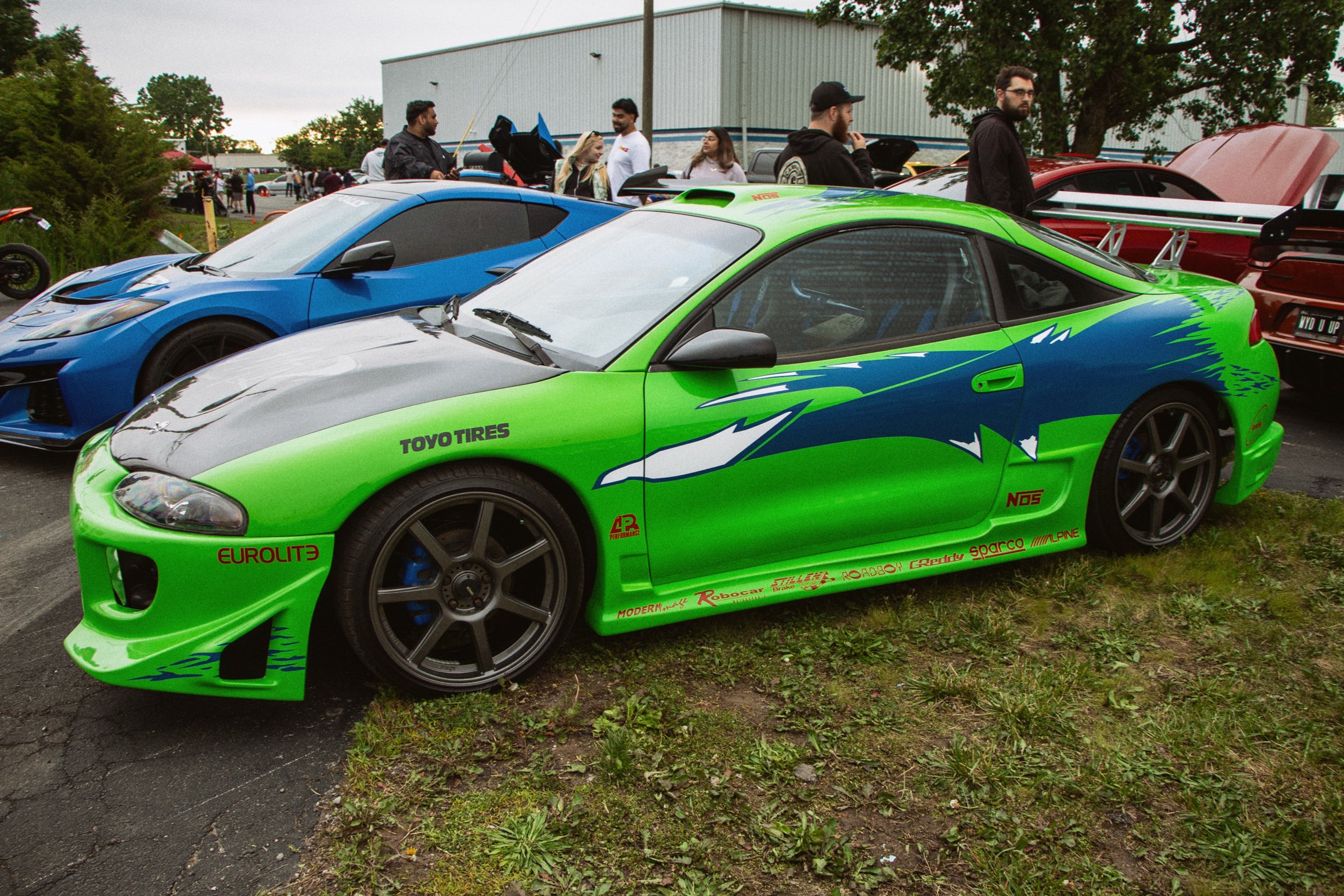 A lineup of colorful race cars at an outdoor car show, with people walking and talking in the background.