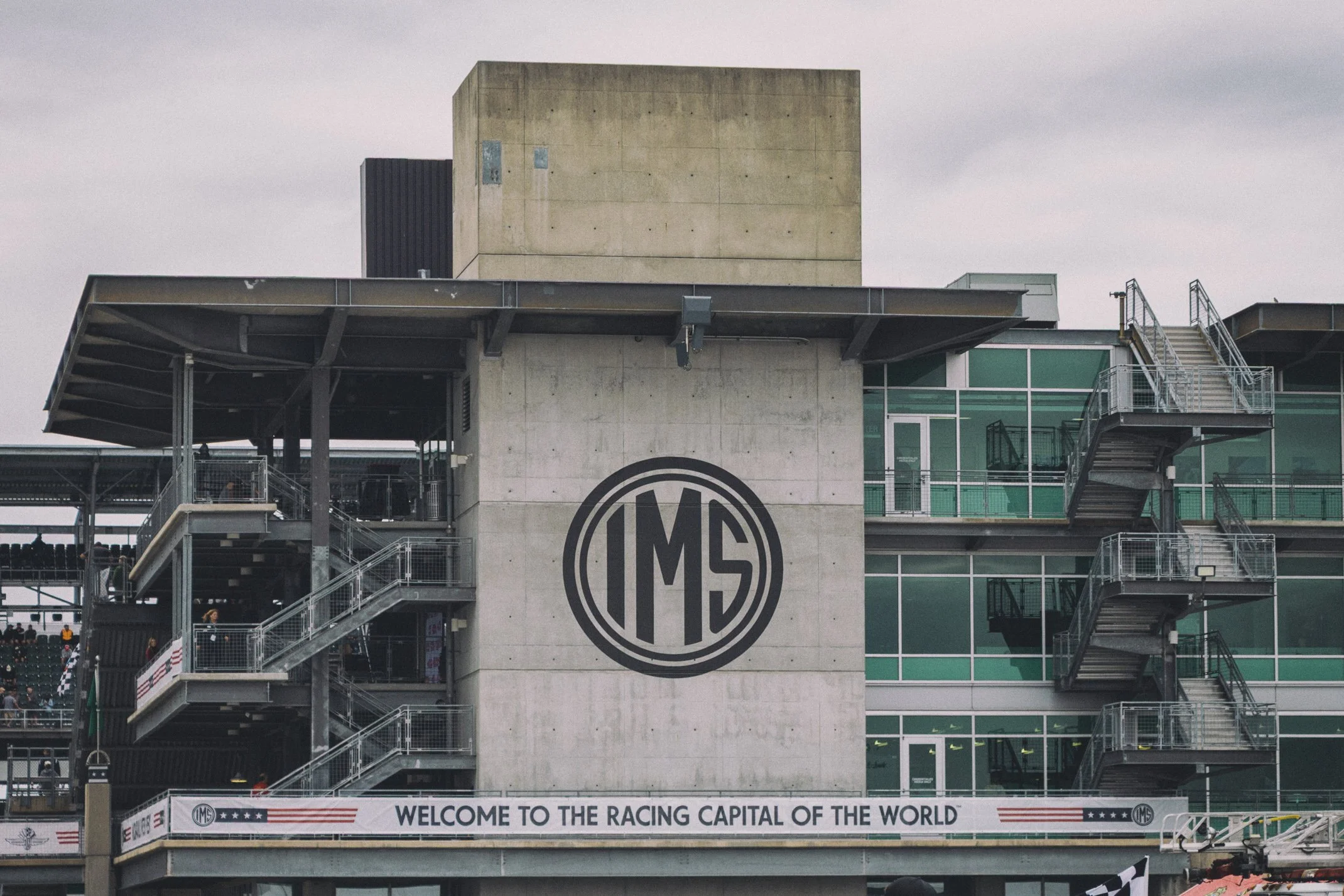 View of a modern racing circuit building with stairs, railings, and signage including IMS logo and a banner that says "Welcome to the racing capital of the world."