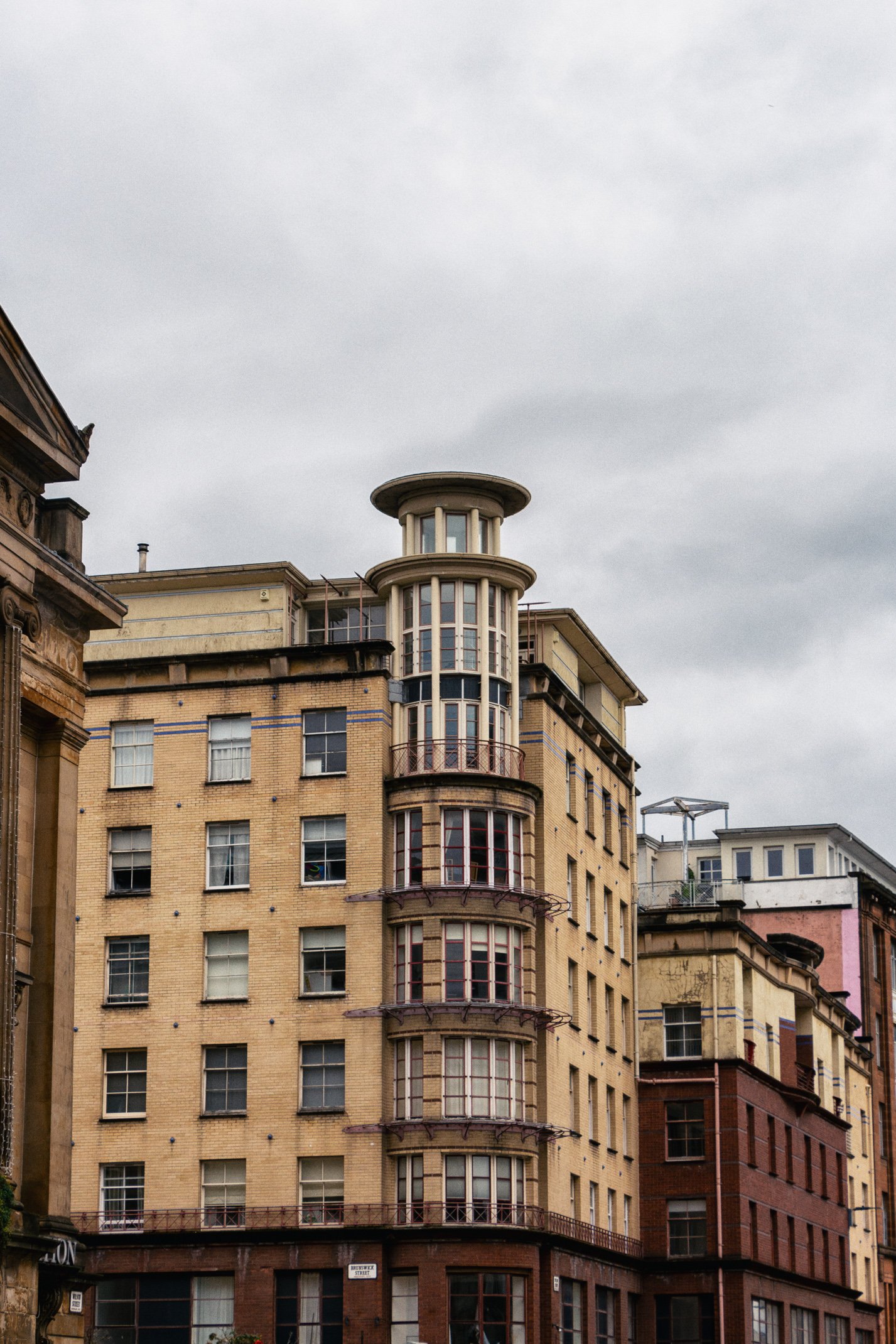 Old brick apartment building with rounded glass corner tower and balconies, under cloudy sky.