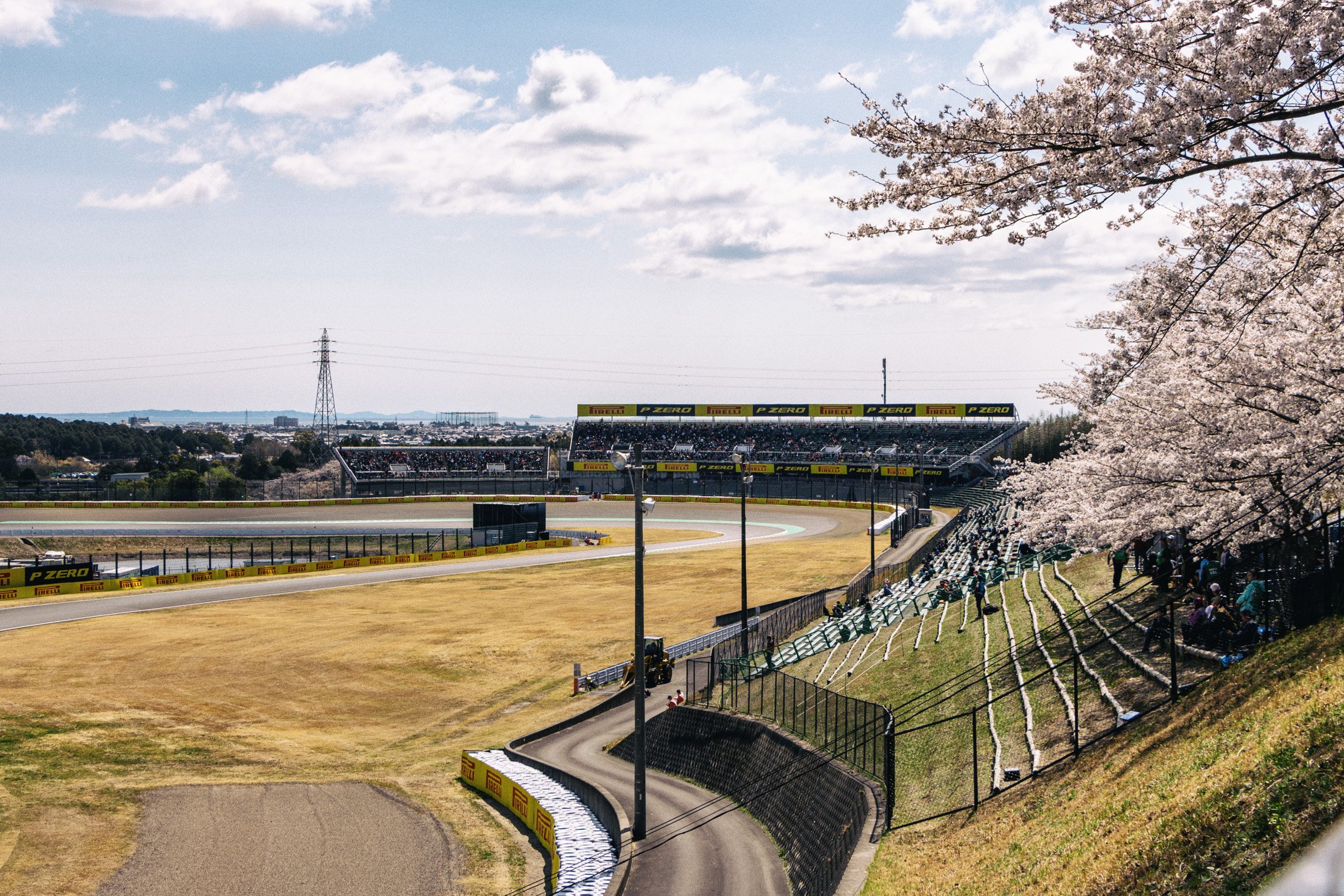 Race track with grandstands, cherry blossom trees, and spectators under a partly cloudy sky.