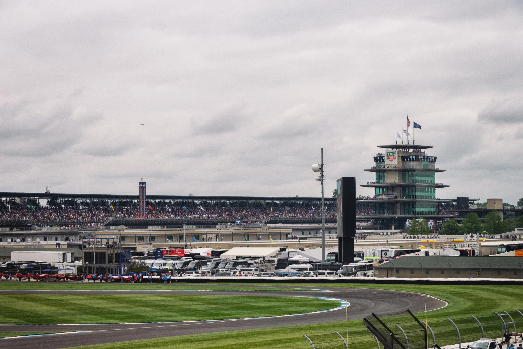 A racing event at a motorsport track with a large crowd in the grandstands. The image shows a multi-story control tower with flags, a logo, and electronic signs, along with a section of the racetrack with a bend in the foreground. The sky is overcast