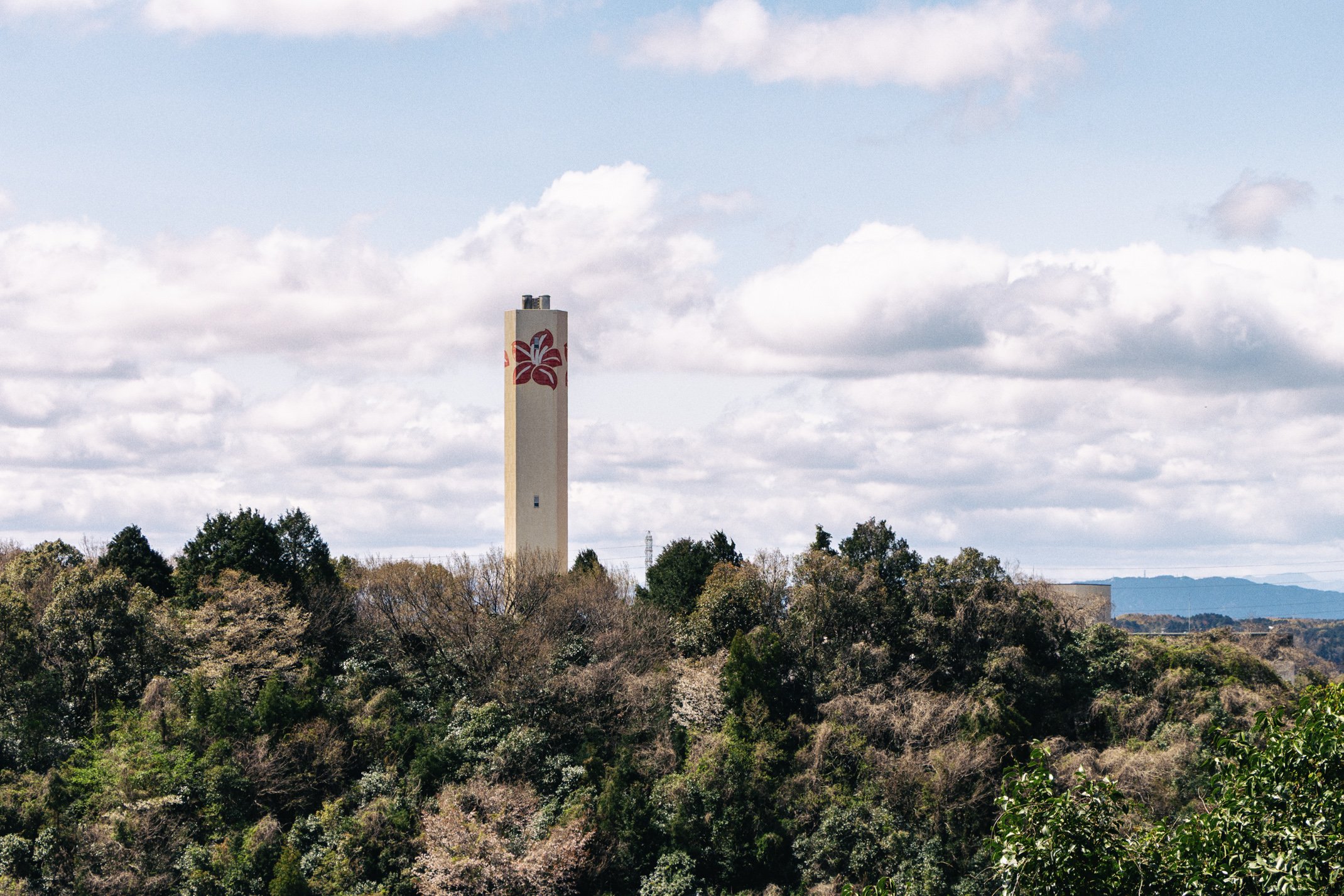 A tall, white tower with a red floral design stands above lush green trees under a partly cloudy sky.