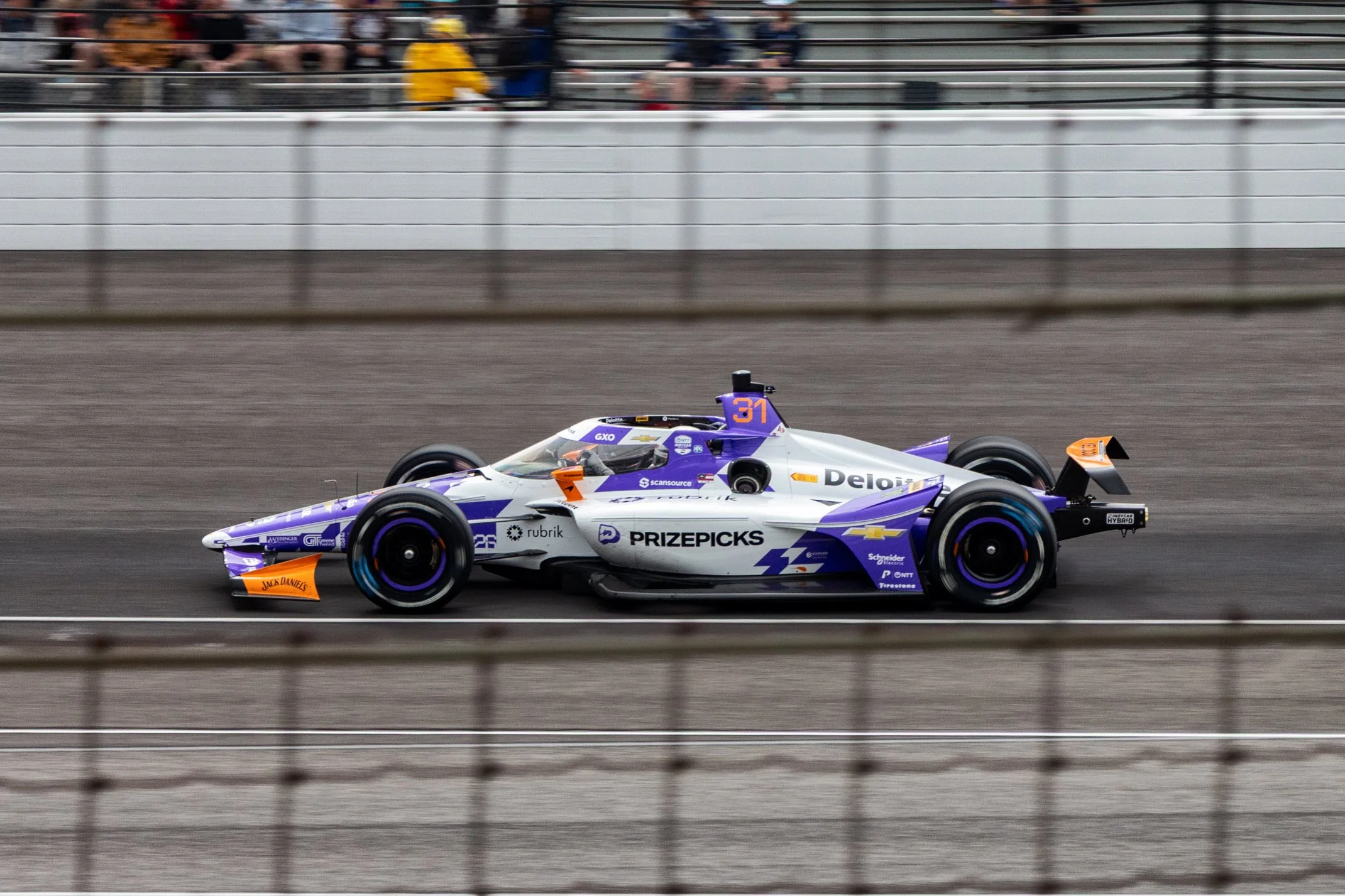 A purple and white IndyCar race car number 31 with various sponsor logos, racing on a track with blurred spectators in the background and a metal fence in the foreground.
