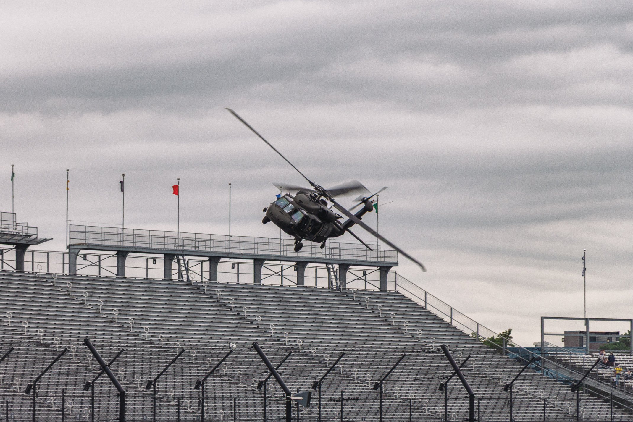 A military helicopter flying low over stadium seating, with flags and cloudy sky in the background.