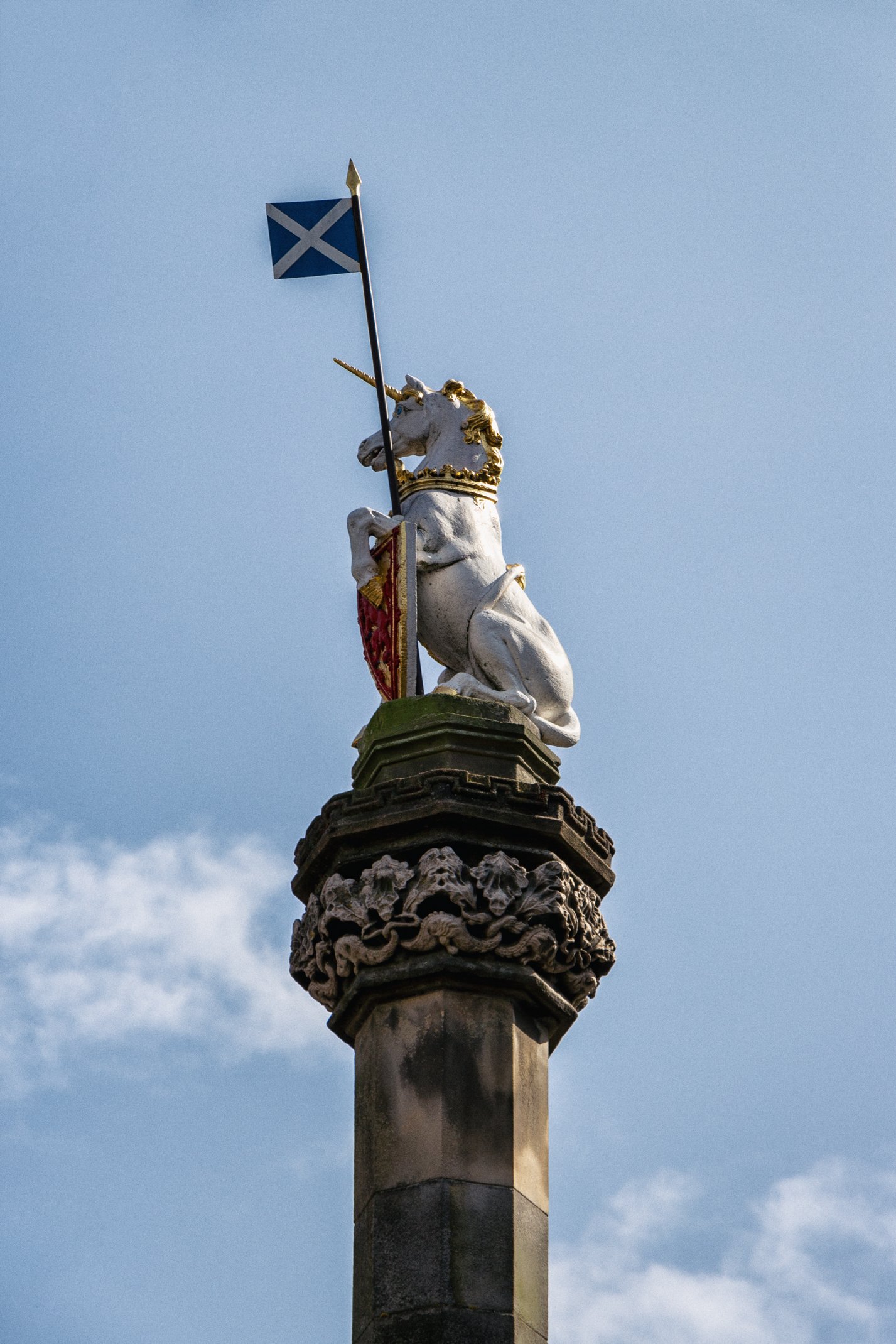 A stone monument featuring a statue of a unicorn wearing gold accents, holding a shield, and flying a Scottish flag on a pole against a cloudy sky.