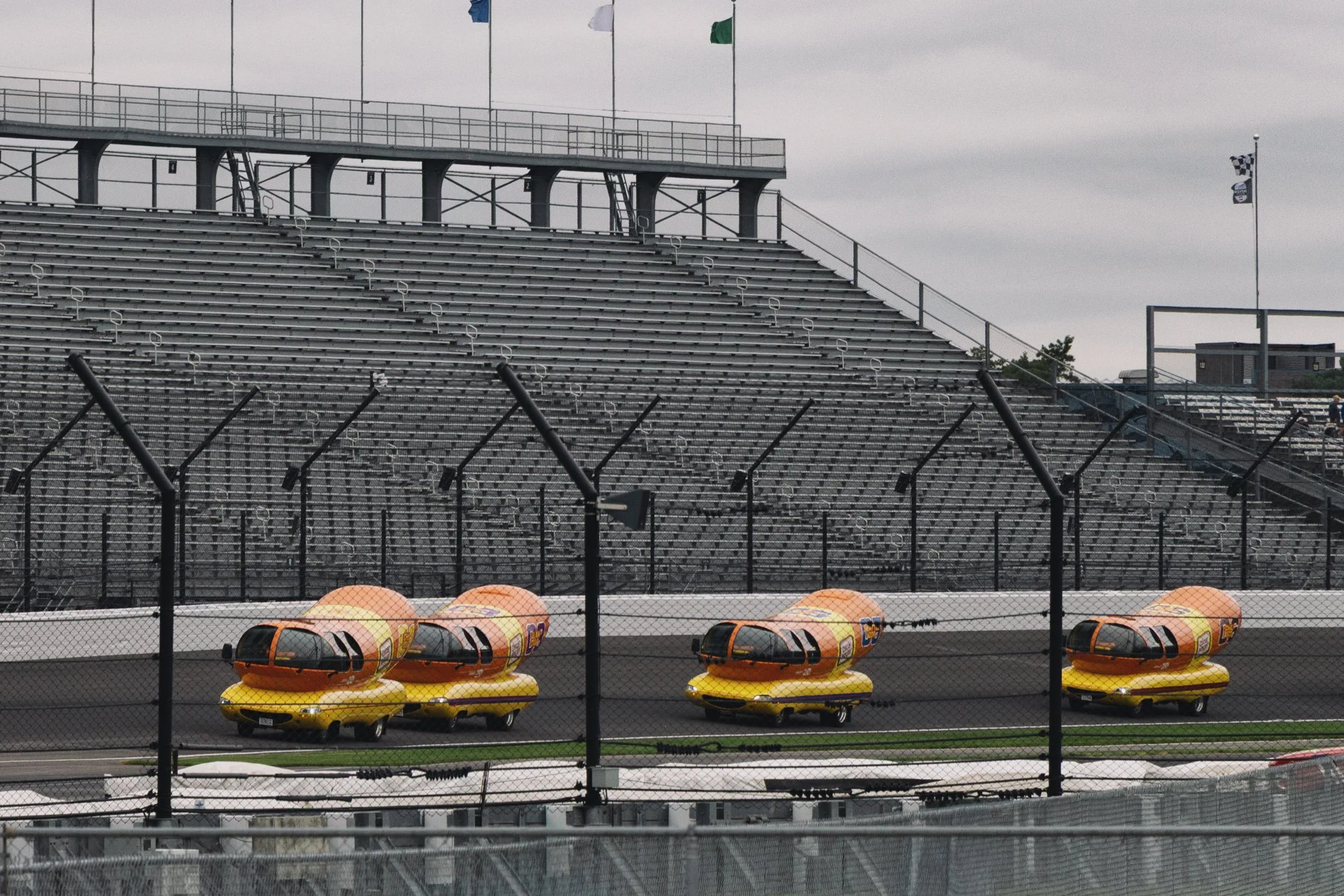 Empty race track with bright yellow race cars in the foreground, sealed with a black fence, and gray stadium seating under an overcast sky.