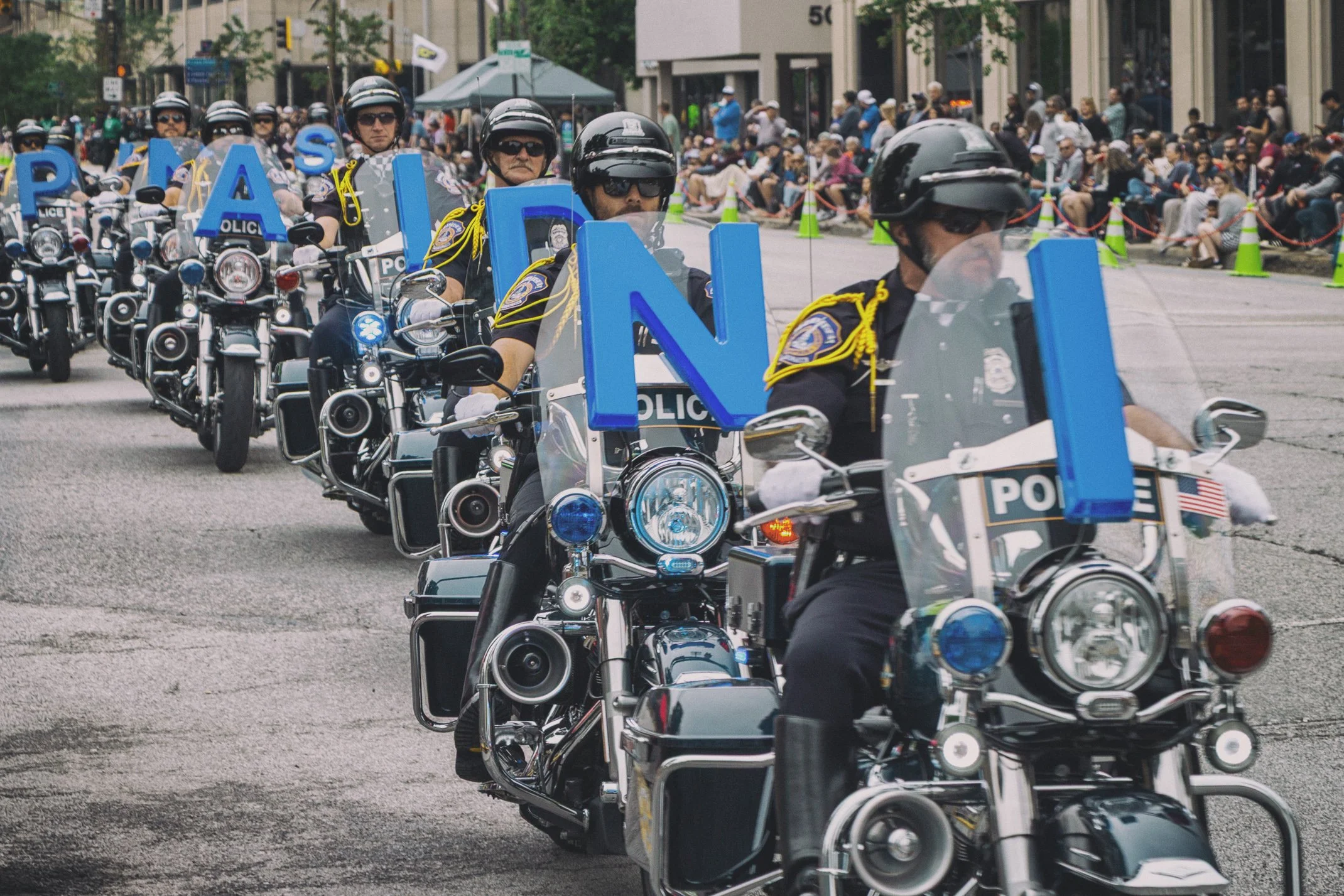 Line of police officers riding motorcycles in a parade, with many spectators seated along the street watching.