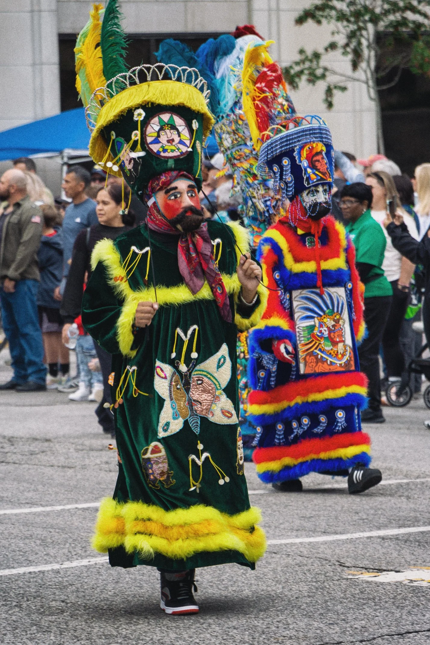 People participating in a cultural parade, wearing traditional colorful costumes and masks, celebrating outdoors with onlookers in the background.