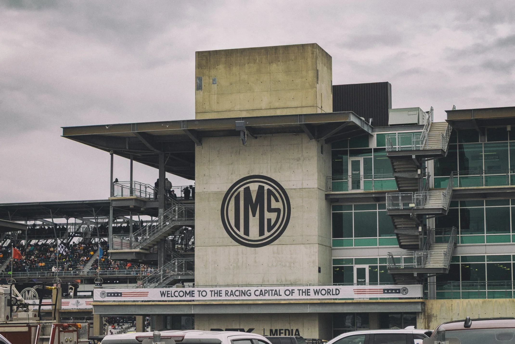Part of a race track with a grandstand filled with spectators, a large building with a concrete facade featuring the IMS logo, and a banner that reads "Welcome to the Racing Capital of the World".