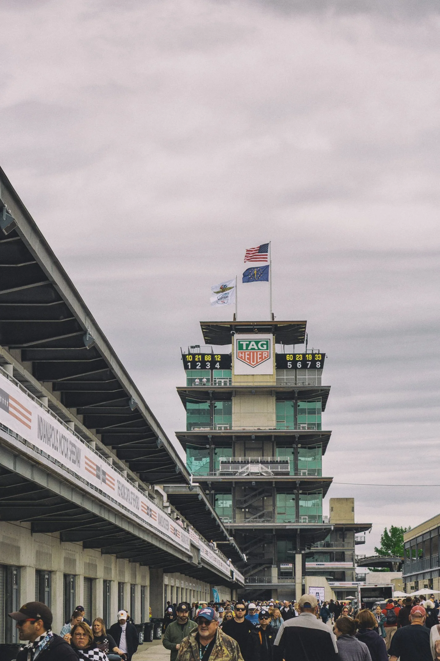 Crowd of people walking at a racing event near the grandstand with flags flying above the building, under a cloudy sky.