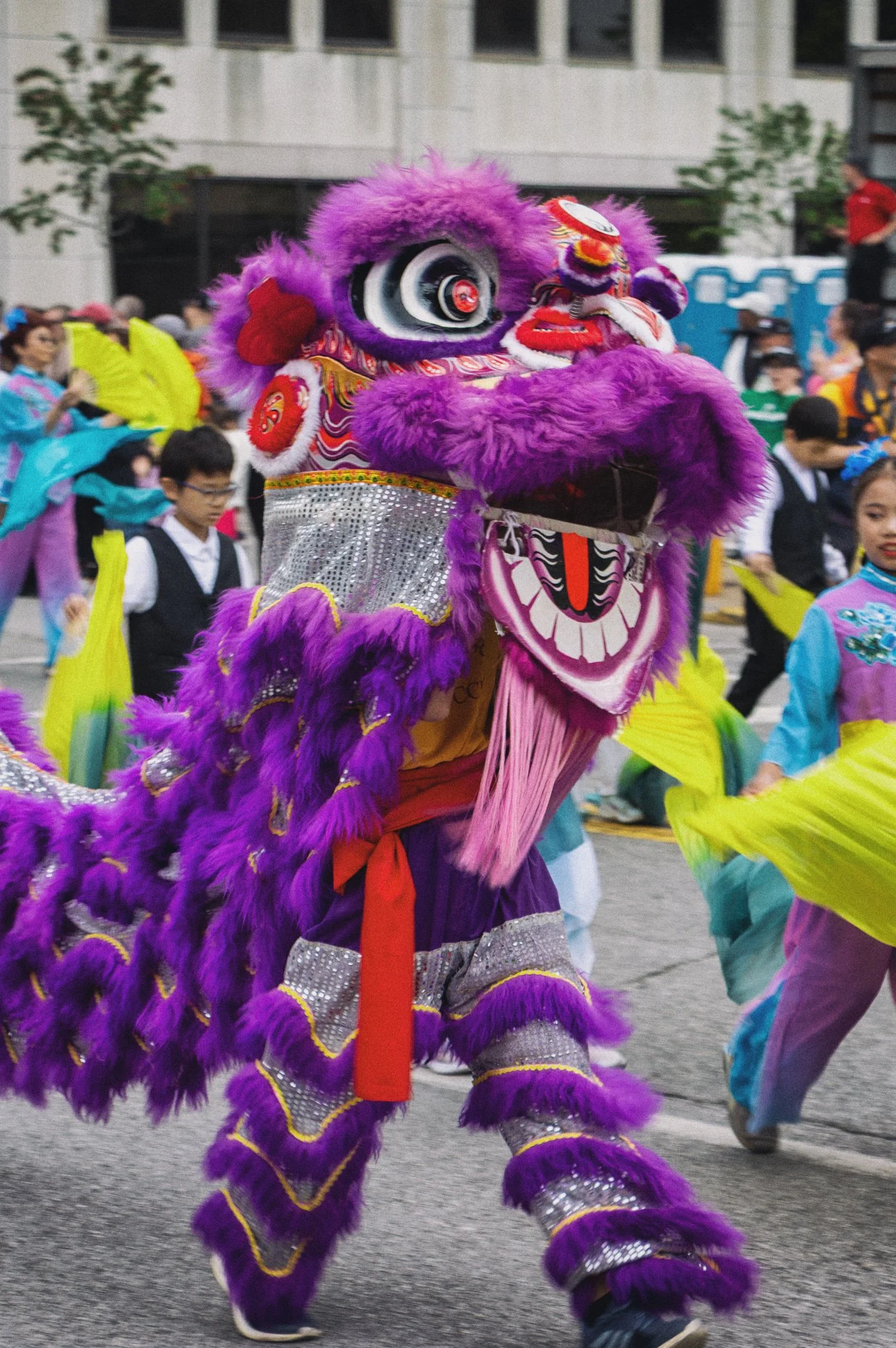 A colorful, traditional lion costume with purple fur, intricate designs, and a large face mask, part of a lion dance parade surrounded by performers and spectators in festive attire.