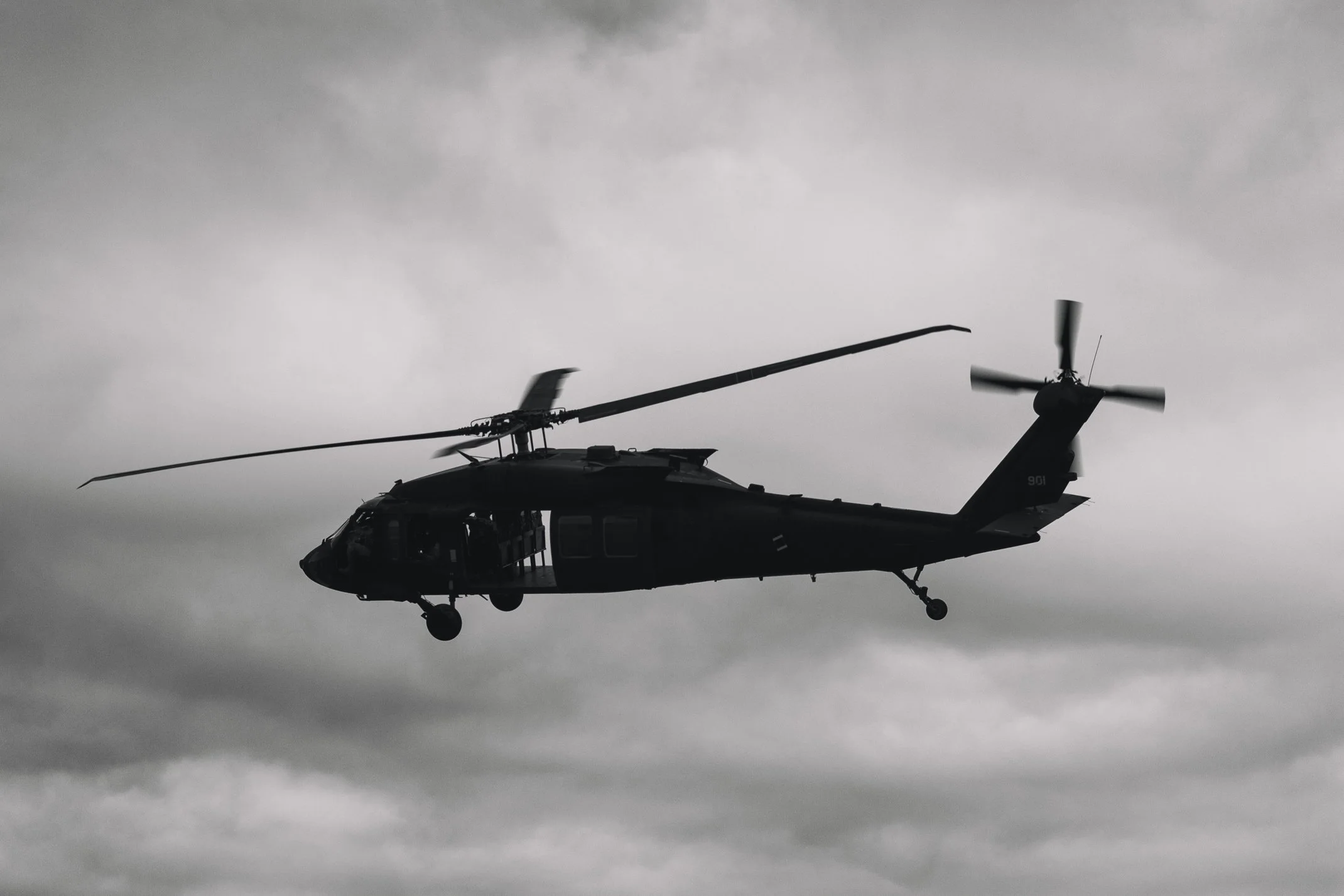 A black military helicopter flying in a cloudy sky.