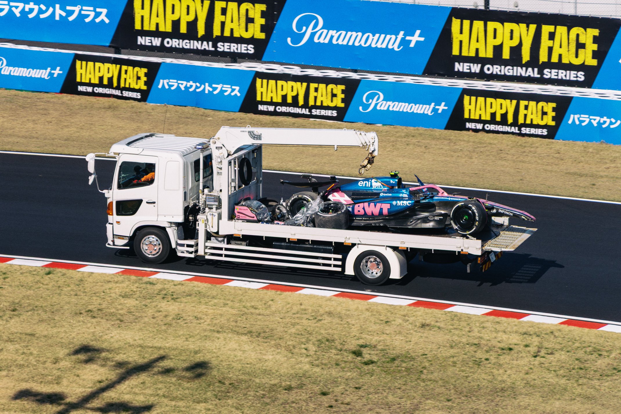 A race car being transported on a flatbed truck at a racing circuit with advertising banners in the background.