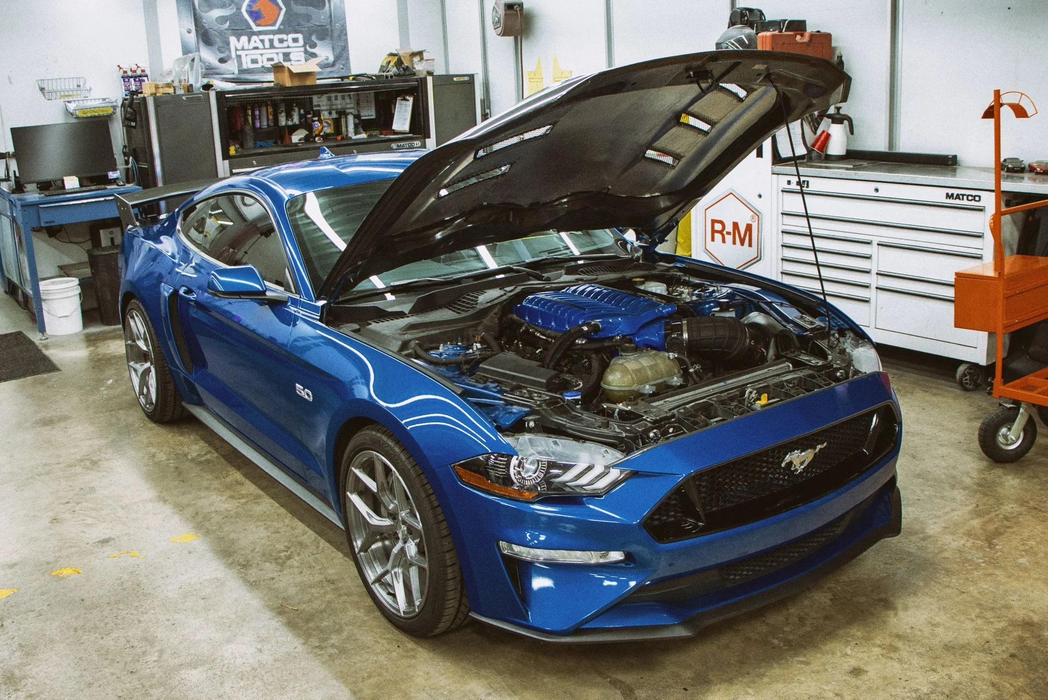 A blue Ford Mustang sports car with its hood open in a garage, revealing the engine. The garage has various tools and storage cabinets, and a workbench with a computer monitor.