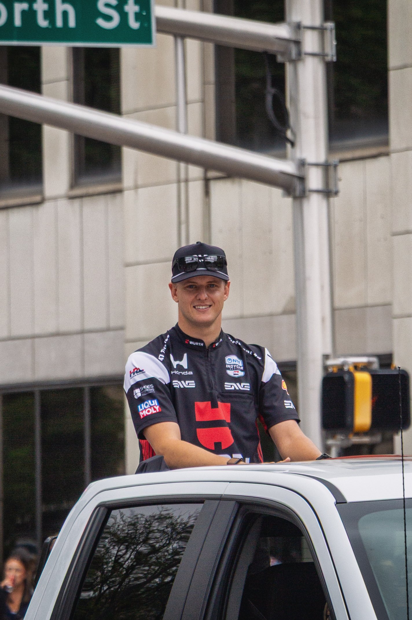 A man wearing a racing suit and sunglasses on his hat, smiling, standing outside behind a white vehicle, with a building, street sign, and traffic light in the background.