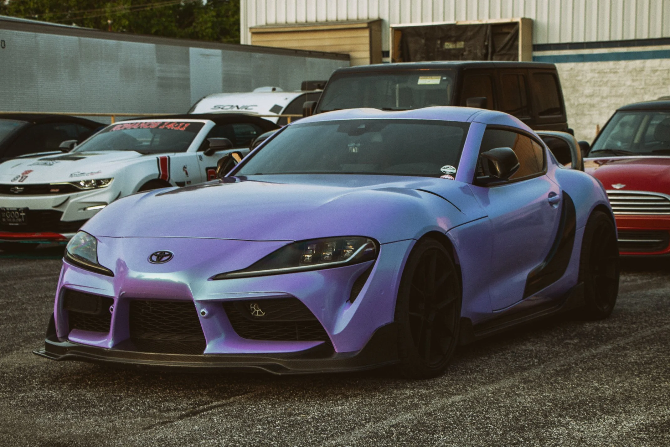 A purple Toyota sports car parked in a lot alongside other cars, including a white racing car with red accents and a red convertible, with industrial buildings in the background.