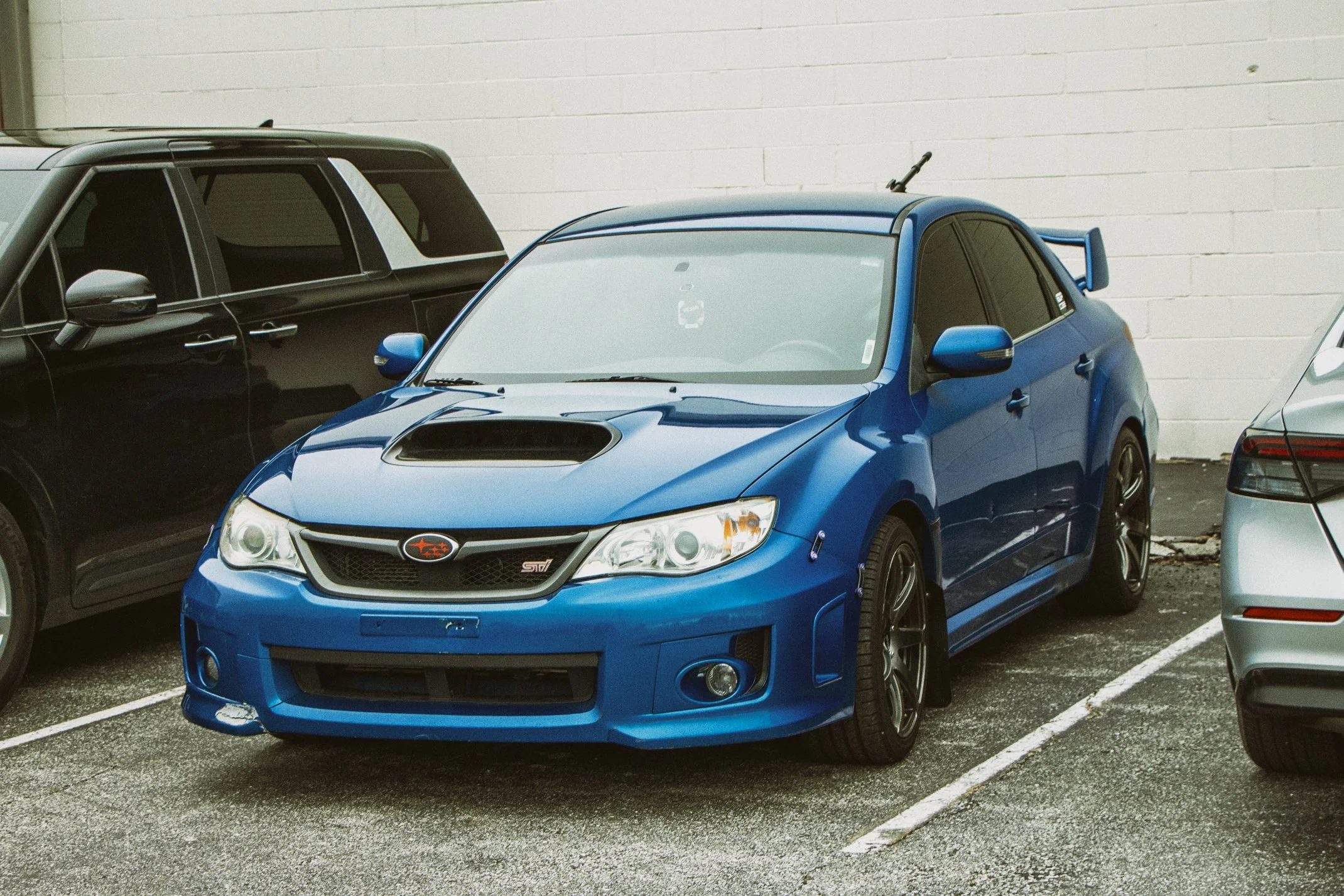 A blue Subaru STI car parked in a parking lot next to a black SUV and a silver car, against a white brick wall.