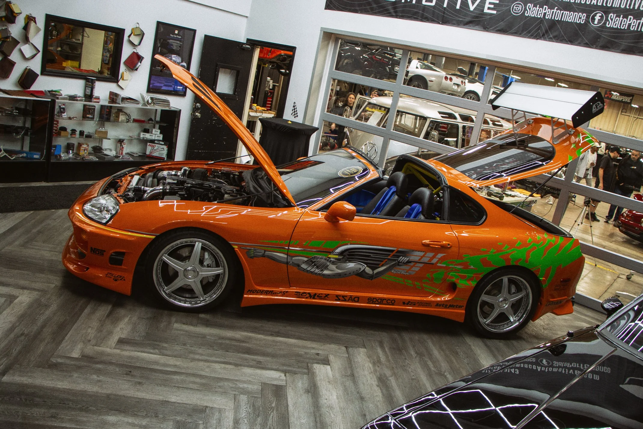 Orange sports car with its hood open displayed at an auto performance shop exhibition, with various car parts and tools in the background.