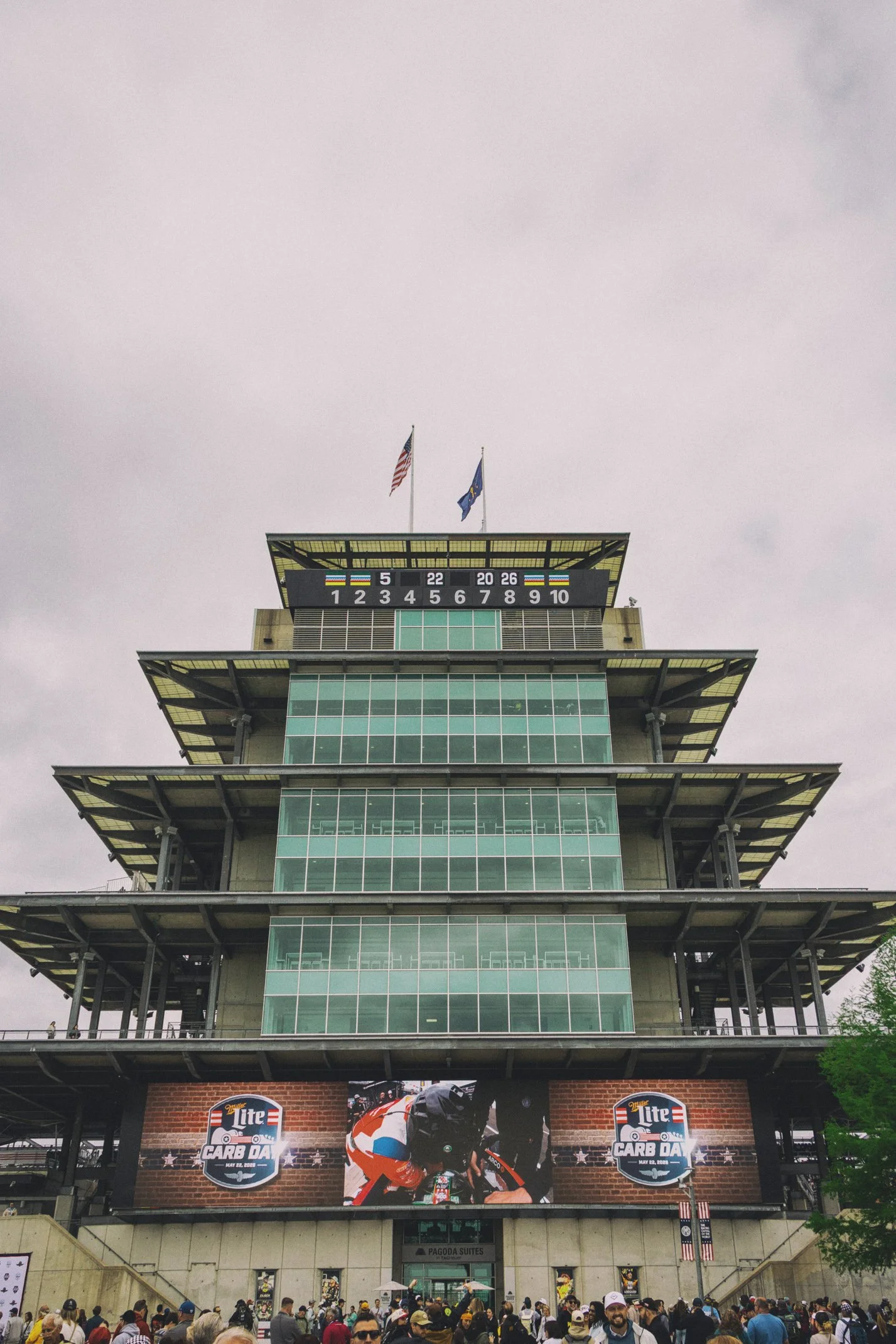 People gathered outside a multi-story building with large glass windows, decorated with banners for a Miller Lite Car Dates event featuring a motorcycle racer and racing bicycle, under an overcast sky.