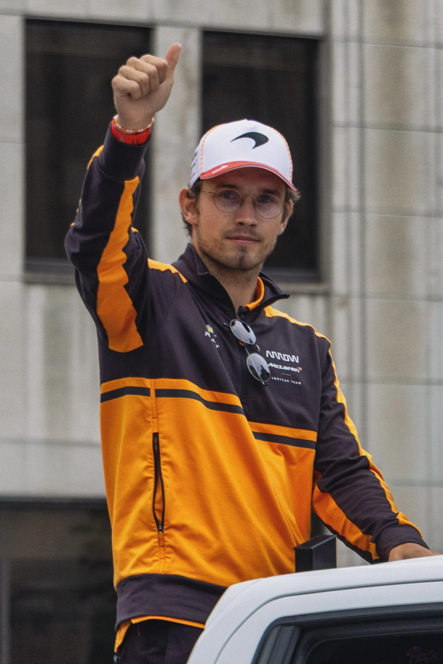 A young man wearing glasses, a white and red cap, and a black and orange jacket, raising his right thumb in a gesture of approval or greeting, standing outdoors near a gray building.