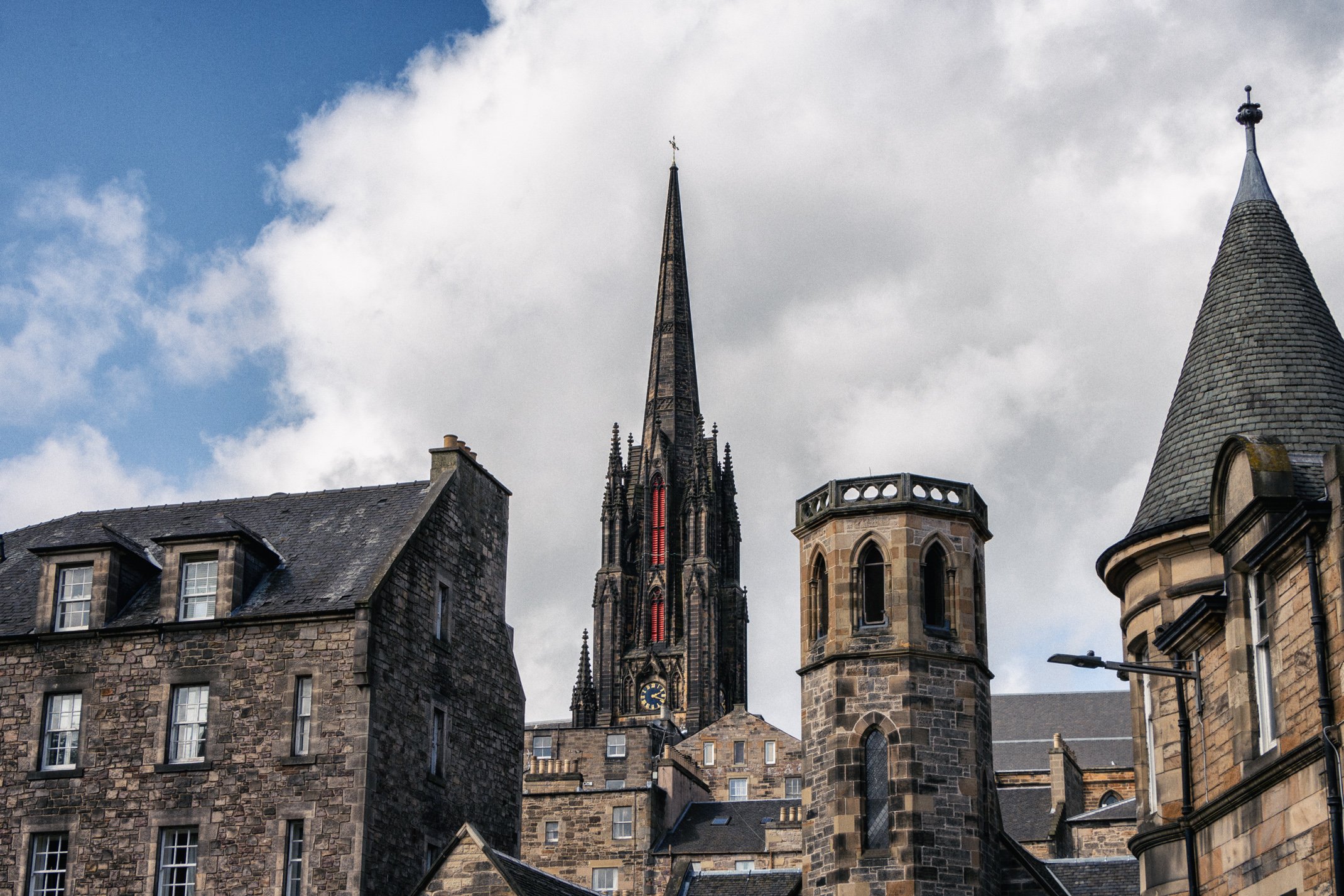 View of old stone buildings and a tall Gothic church steeple against a partly cloudy sky.