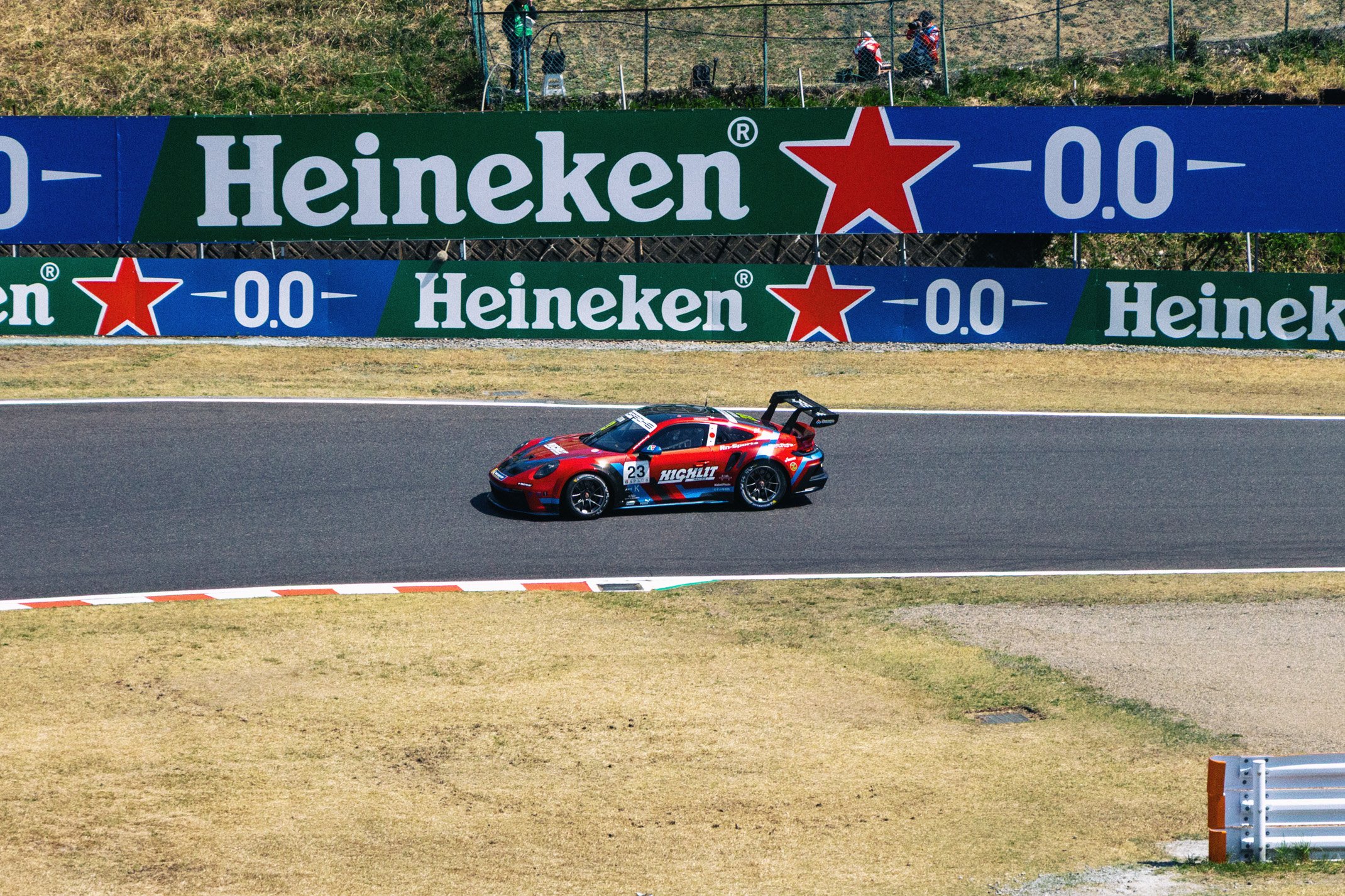 A red race car with the number 23 driving on a racetrack, surrounded by advertising banners for Heineken and other sponsors, with a grassy area and a few spectators in the background.