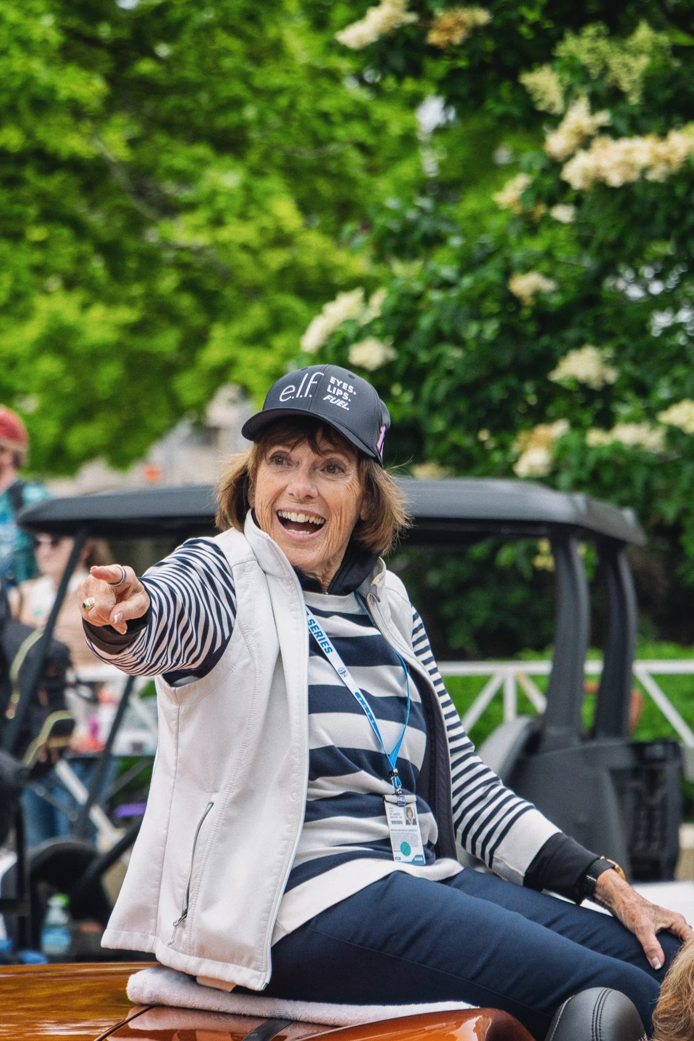 A smiling woman with short brown hair wearing a black cap with white text, a striped navy and white shirt, and a white vest, sitting on the back of a vehicle outdoors during daytime, surrounded by trees and other people.