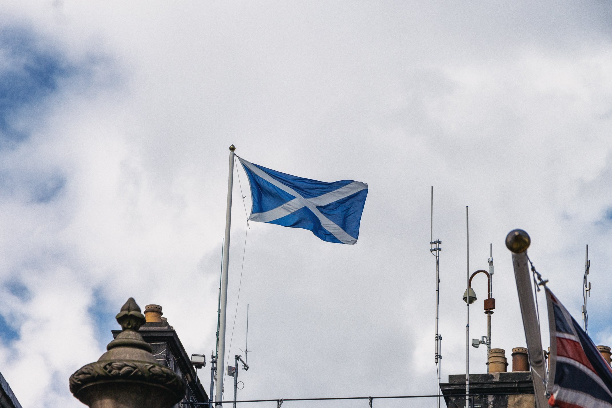Scottish flag flying on a flagpole against a cloudy sky, with antennas and part of a building rooftop visible.