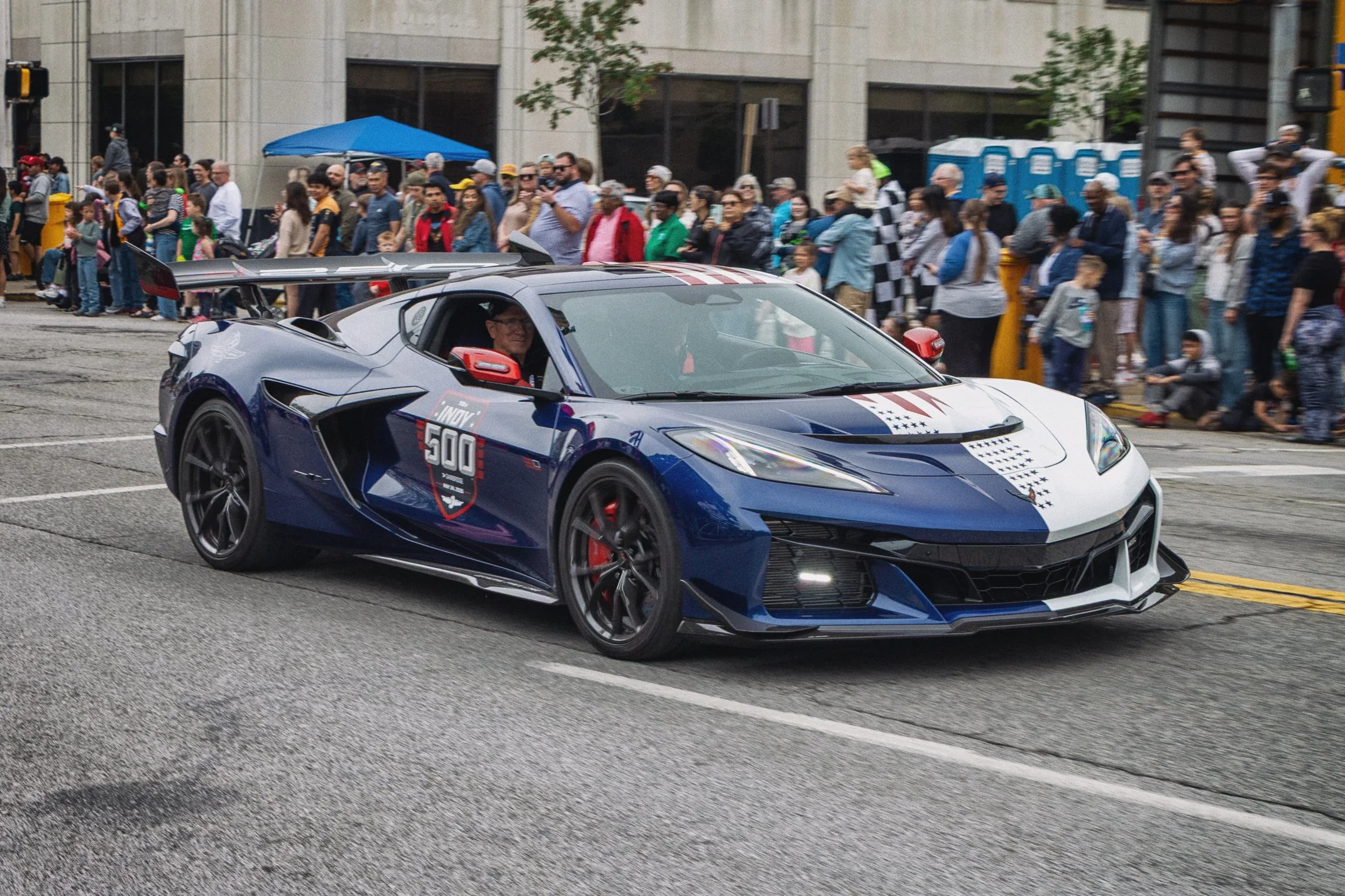 A race car with a driver inside, passing through a crowd of onlookers on a city street during an event, with portable toilets and a tent in the background.