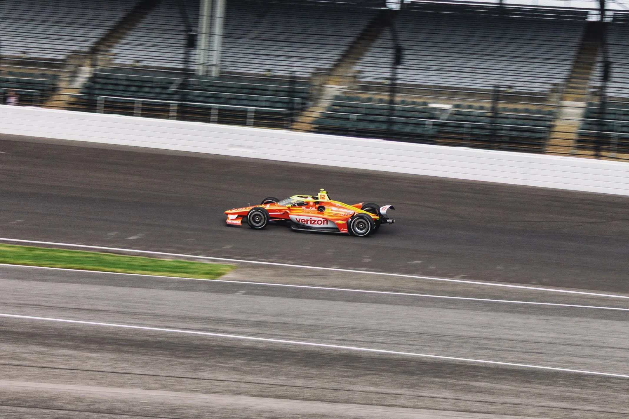 A race car with Verizon sponsorship speeds along a racetrack, with empty grandstands in the background.
