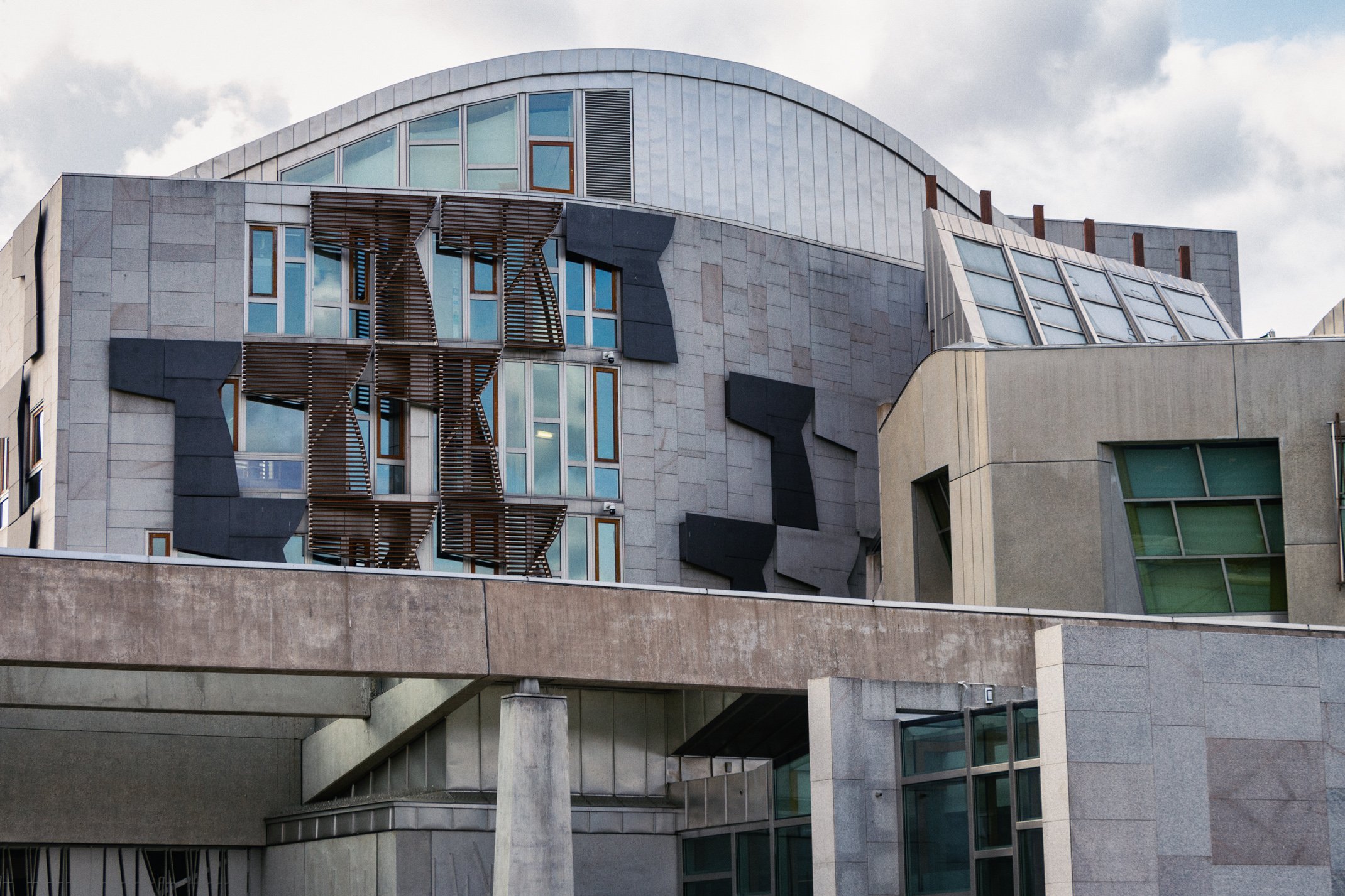 Modern multi-story building with geometric design, featuring large windows, black panel accents, and wooden sunshades, under a partly cloudy sky.