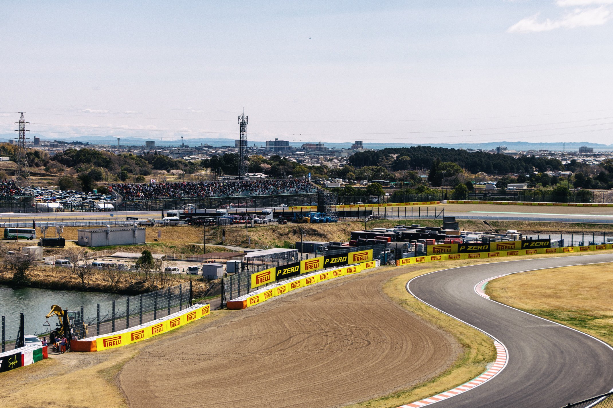 A view of a race track with advertisements from Pirelli and P Zero along the edge, with a turn in the track and a large crowd of spectators in the stands in the background. There are hills and buildings in the distant background under a partly cloudy