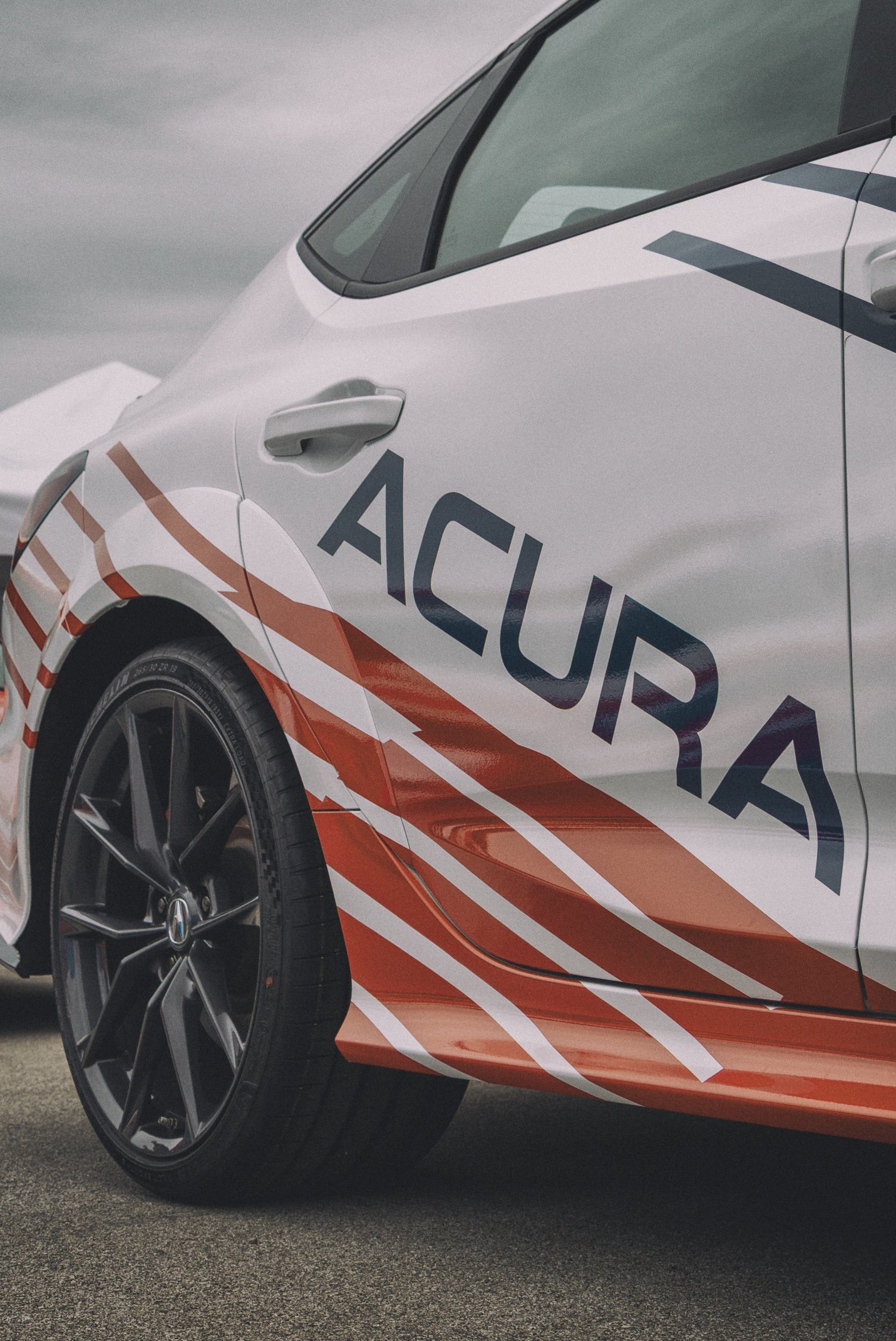 Close-up of a car with the words 'ACURA' on the side in a style resembling a racing stripe, featuring a black wheel with silver accents and a black tire, parked on asphalt with a cloudy sky in the background.