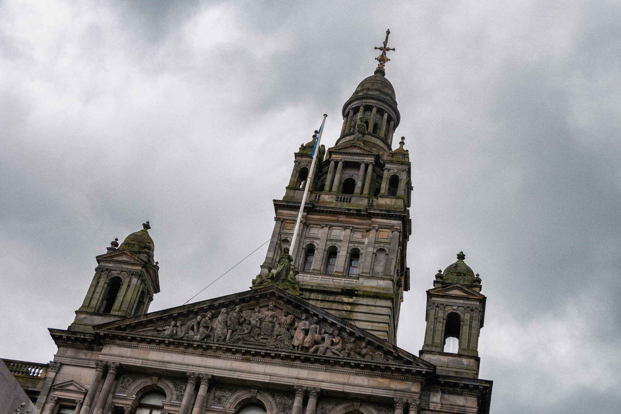 A historic building with a tall clock tower, classical architectural details, sculptures, and a metallic cross at the top, under a cloudy sky.